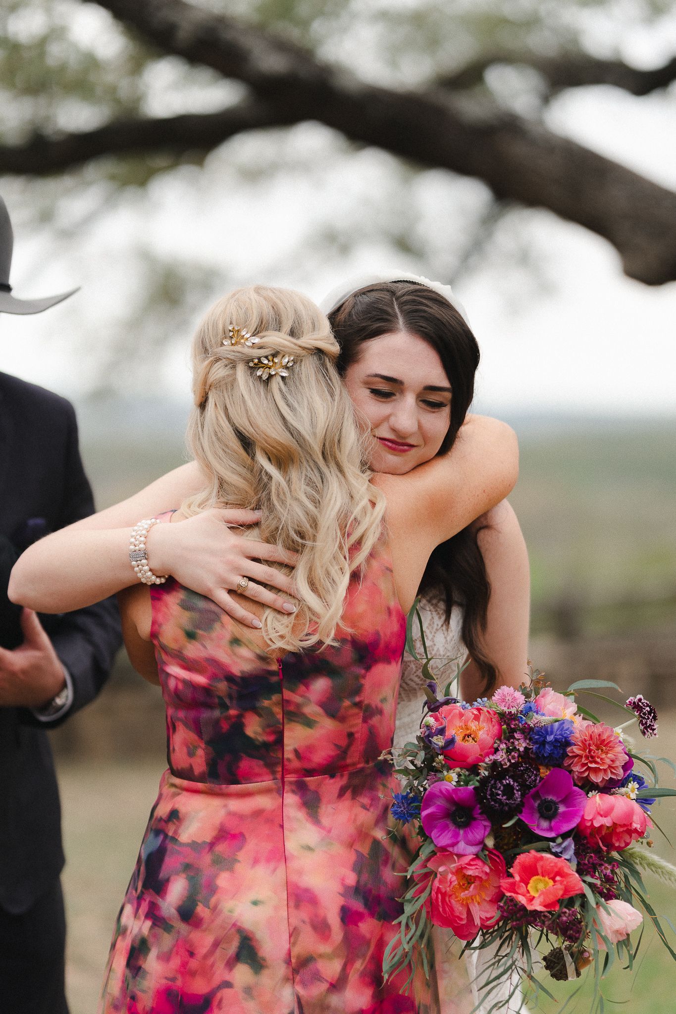 Bride holding colorful bouquet embraces woman in floral pink dress at outdoor wedding ceremony.