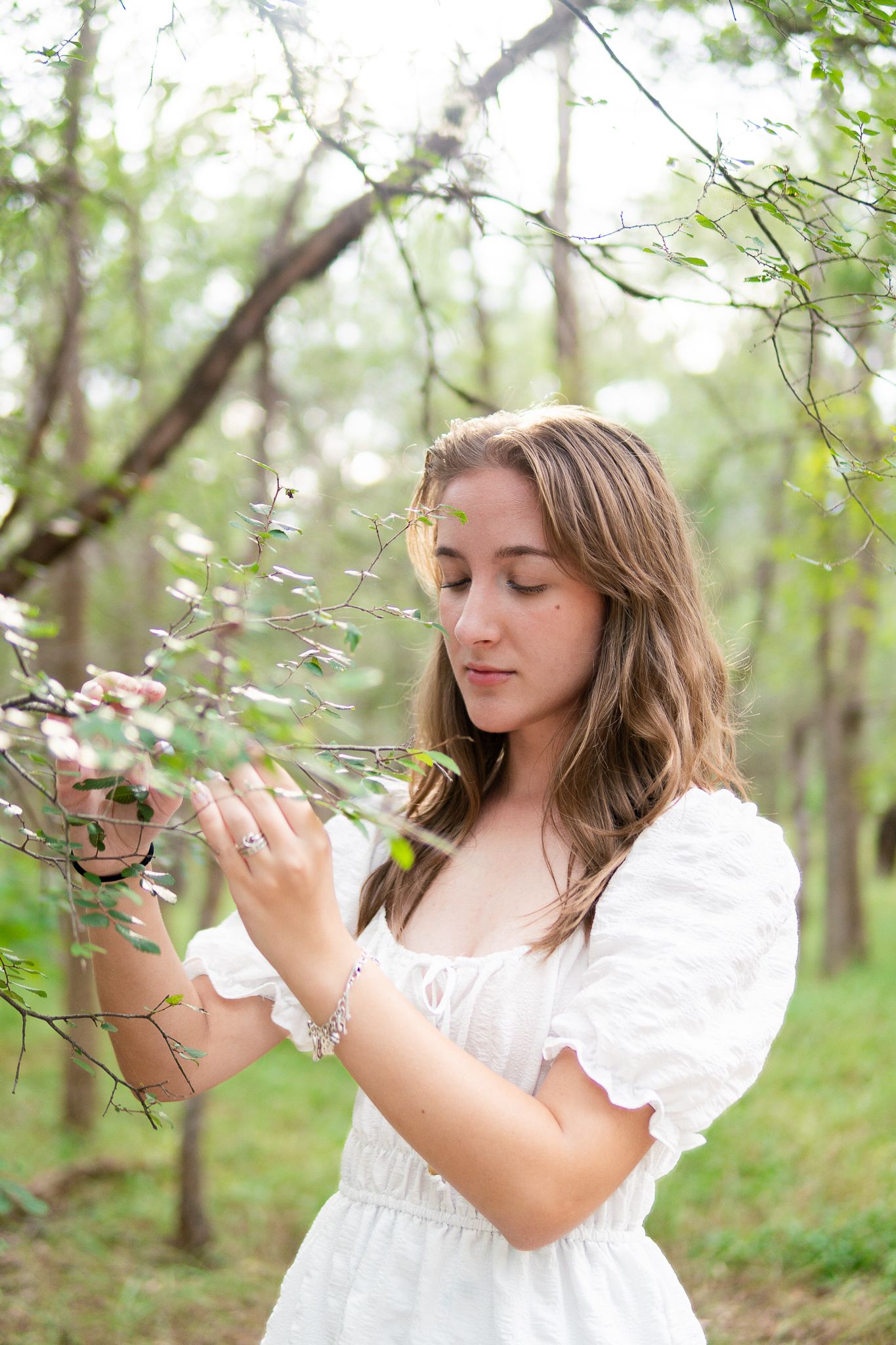 A person in a white dress gently touches spring blossoms in a serene forest setting.