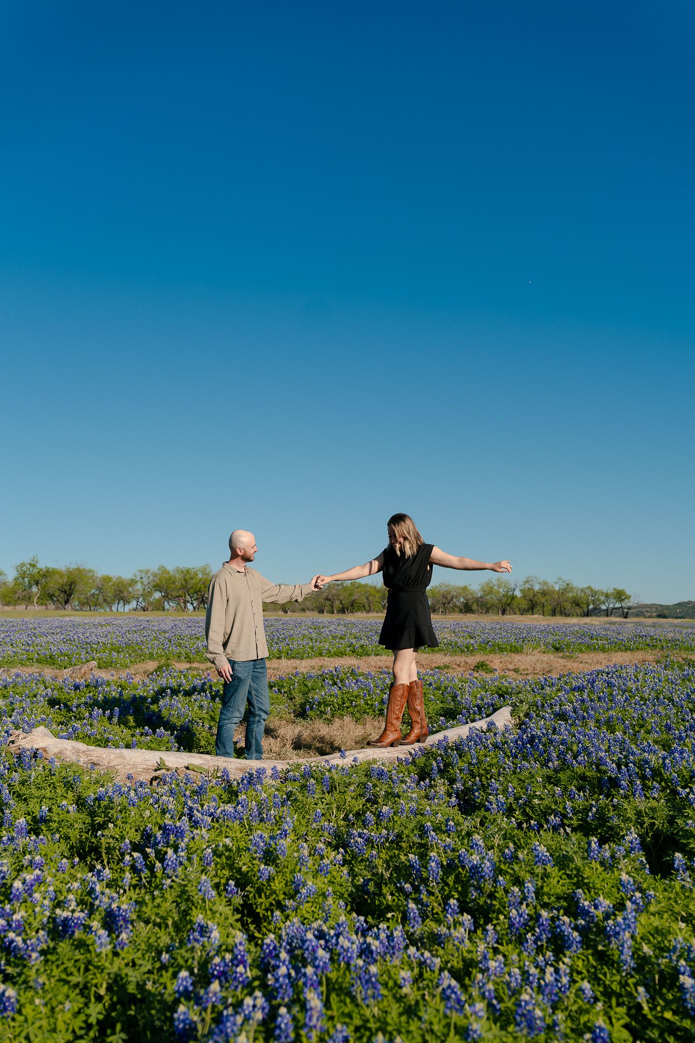 Three people stand with outstretched arms in a vibrant field of blooming Texas bluebonnets under a clear blue sky.