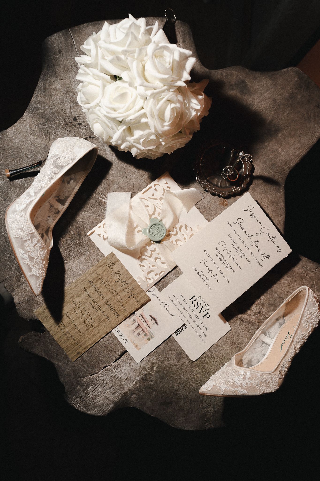 Flat lay of white lace bridal heels, a white rose bouquet, and an elegant wedding invitation suite with RSVP card and wax seal.