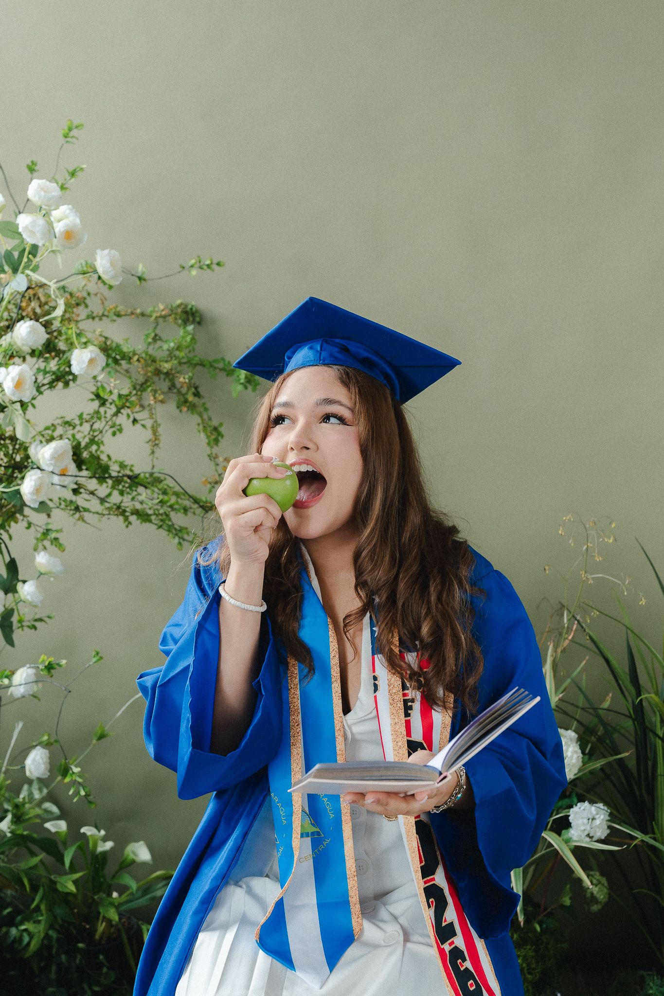 Female graduate in blue cap and gown eating a green apple while holding a book, surrounded by white flowers.