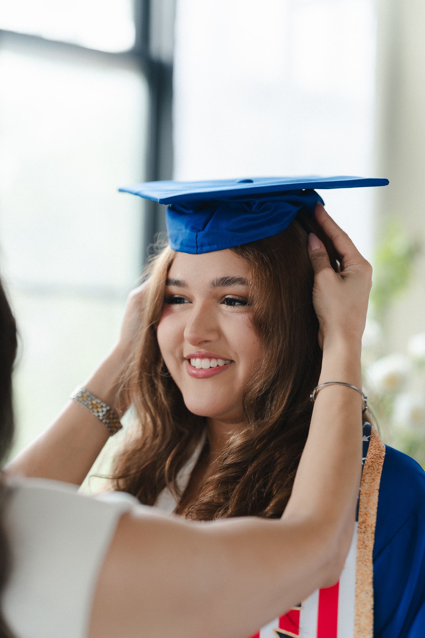 Smiling young woman in blue graduation cap and gown having her mortarboard adjusted at graduation ceremony.