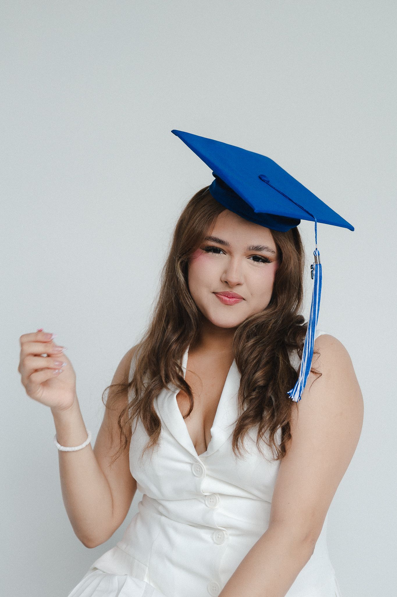 Young woman in white dress wearing blue graduation cap smiling confidently against light background.