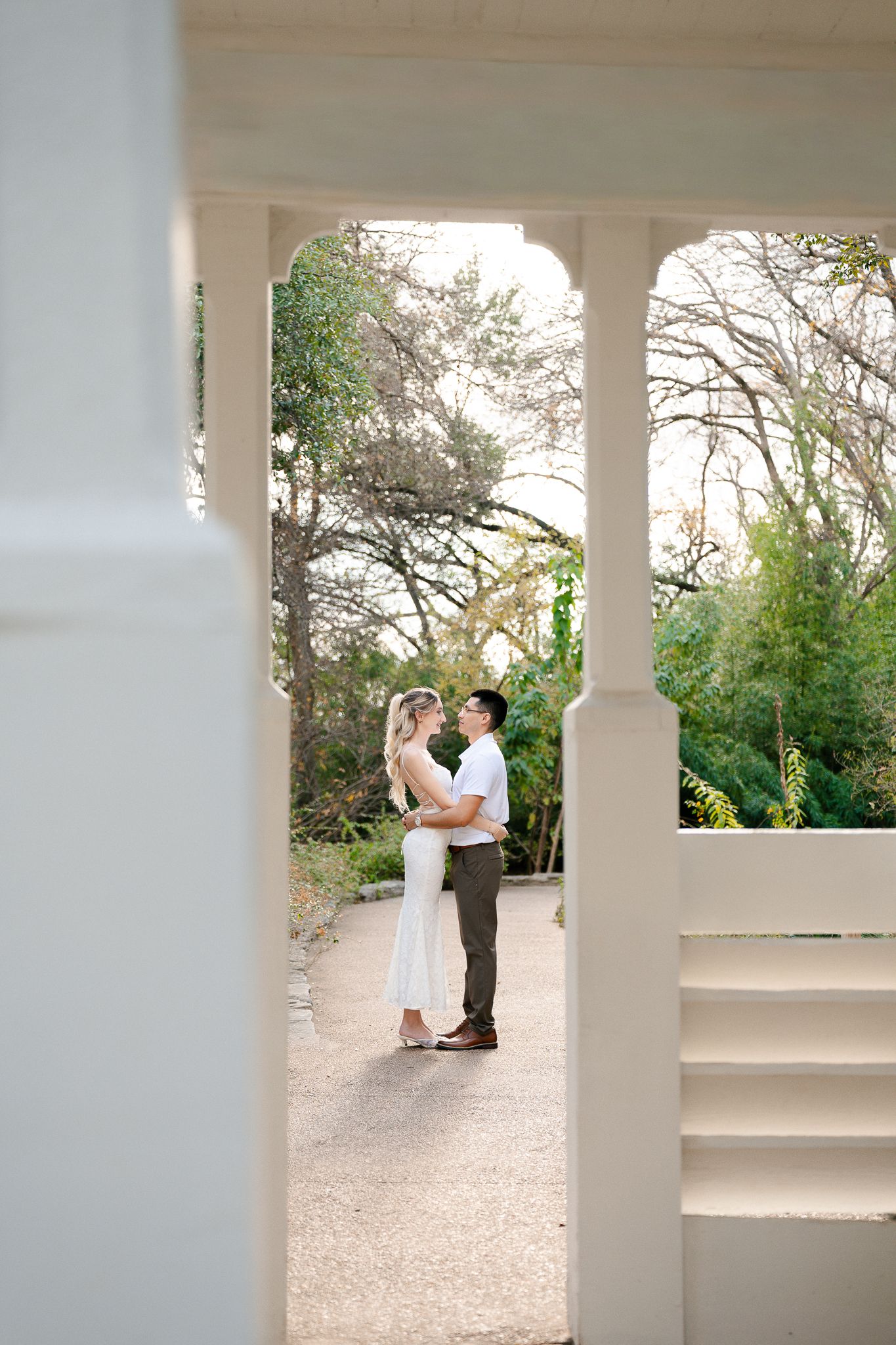 A romantic couple embraces on a garden path framed by white pillars and lush greenery on a bright sunny day.