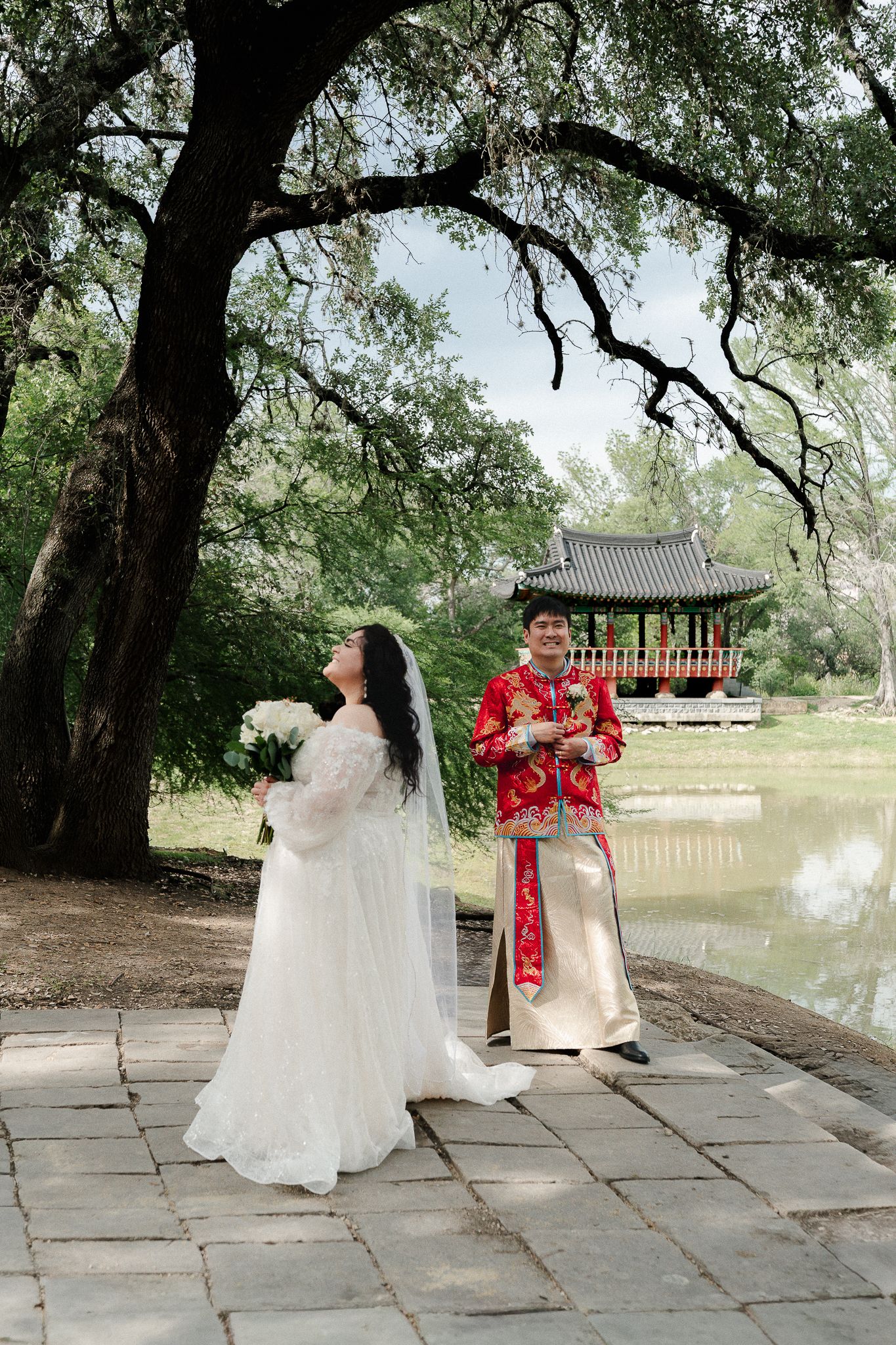 Bride in white gown and groom in red traditional Chinese attire pose near a pagoda by a serene pond in a lush park.
