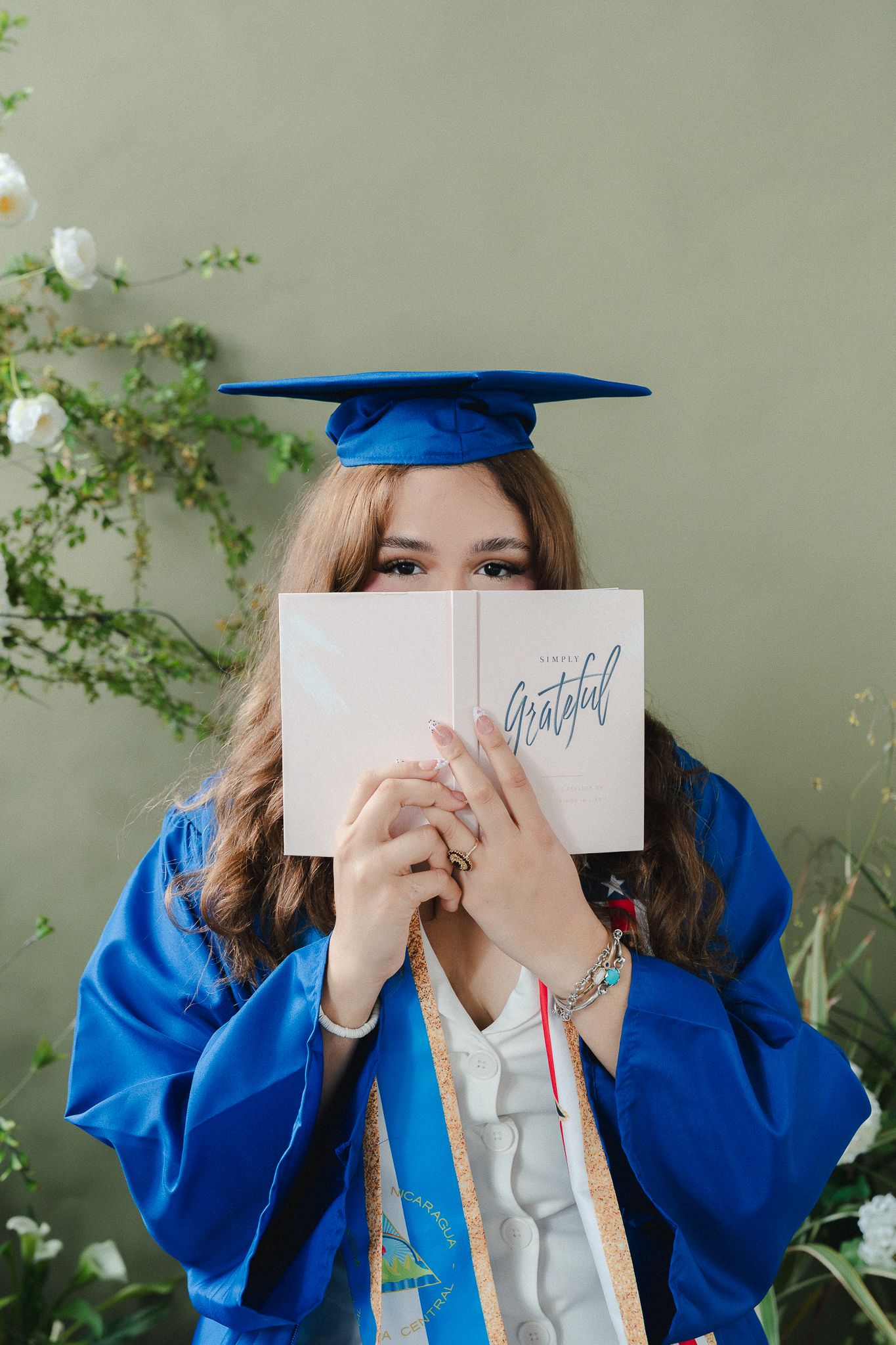 Graduate in blue cap and gown holds a 'Grateful' book, posing among white flowers against a green wall.