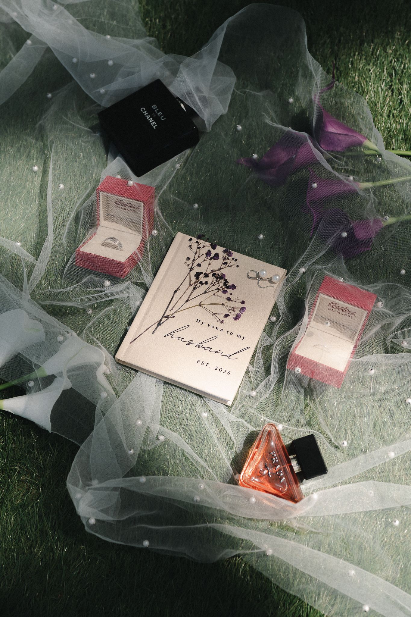Wedding flat lay with veil, ring boxes, perfume, and a husband journal on green grass with purple flowers.