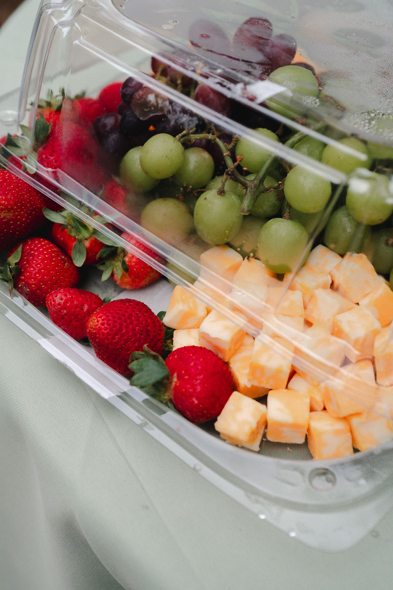 Clear plastic container filled with fresh strawberries, green grapes, red grapes, and cubed cantaloupe melon pieces.