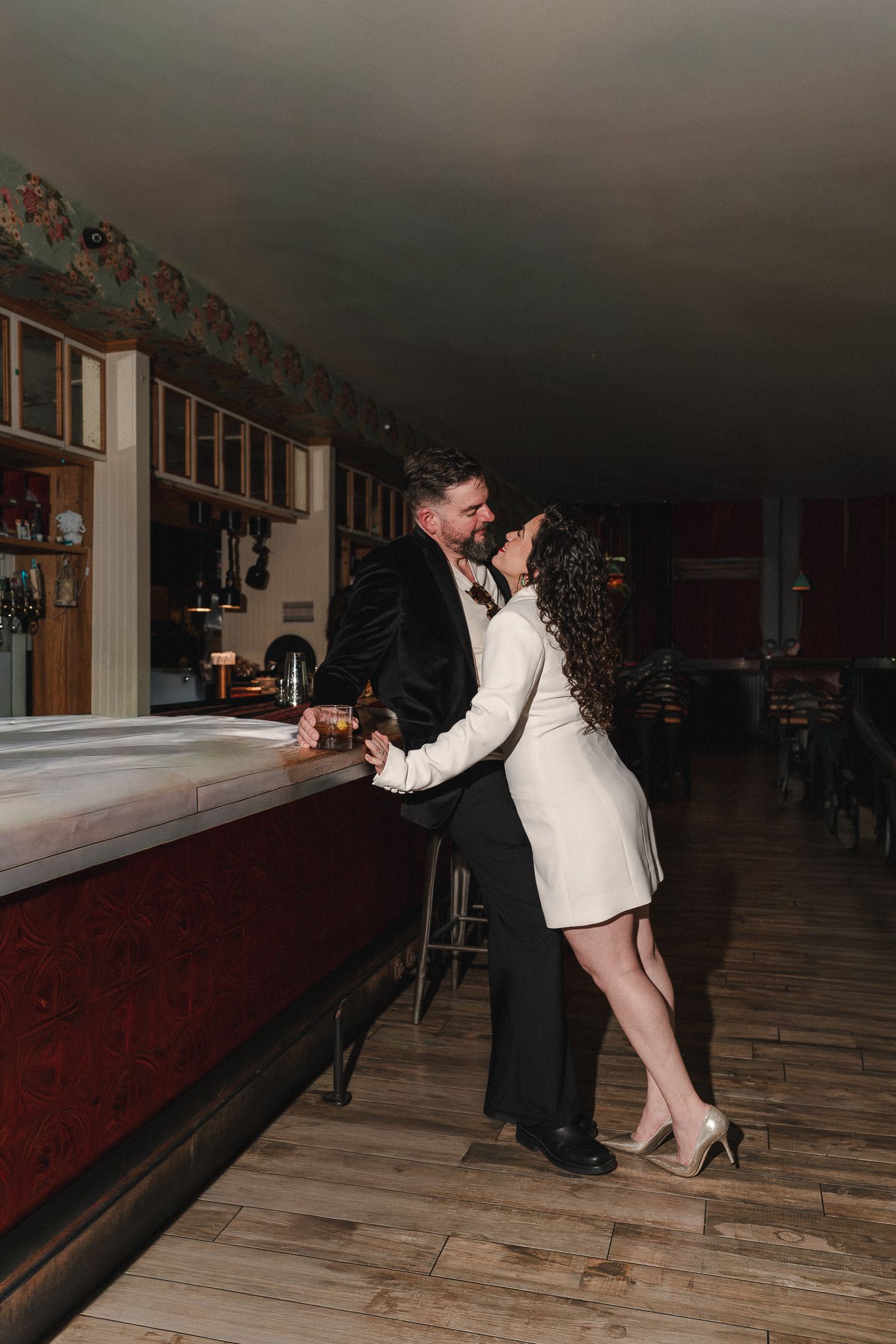 Couple sharing a romantic kiss at a dimly lit bar, dressed in elegant wedding attire, standing on hardwood floors.
