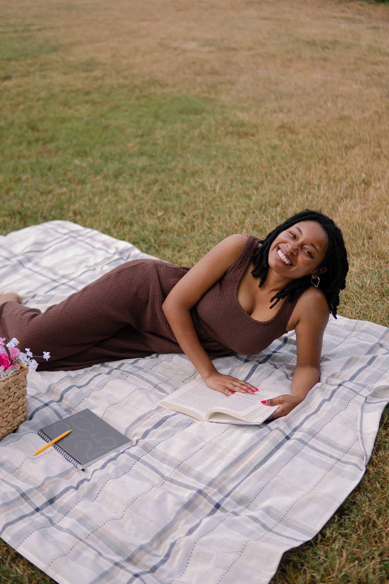 A person relaxes on a blanket outdoors while reading and studying with books and electronic devices nearby.
