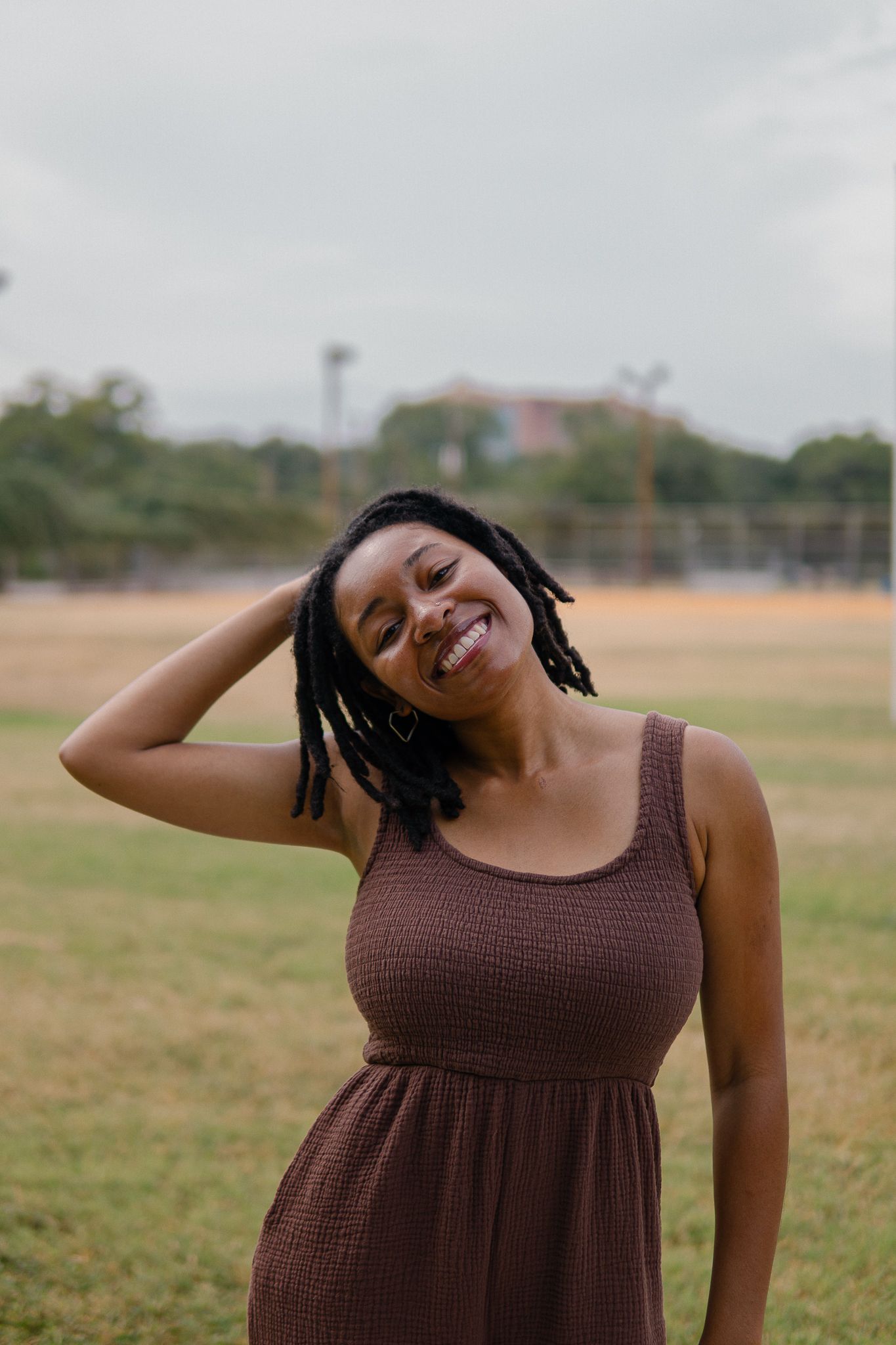 A person in a brown sleeveless dress poses expressively in an outdoor field setting.