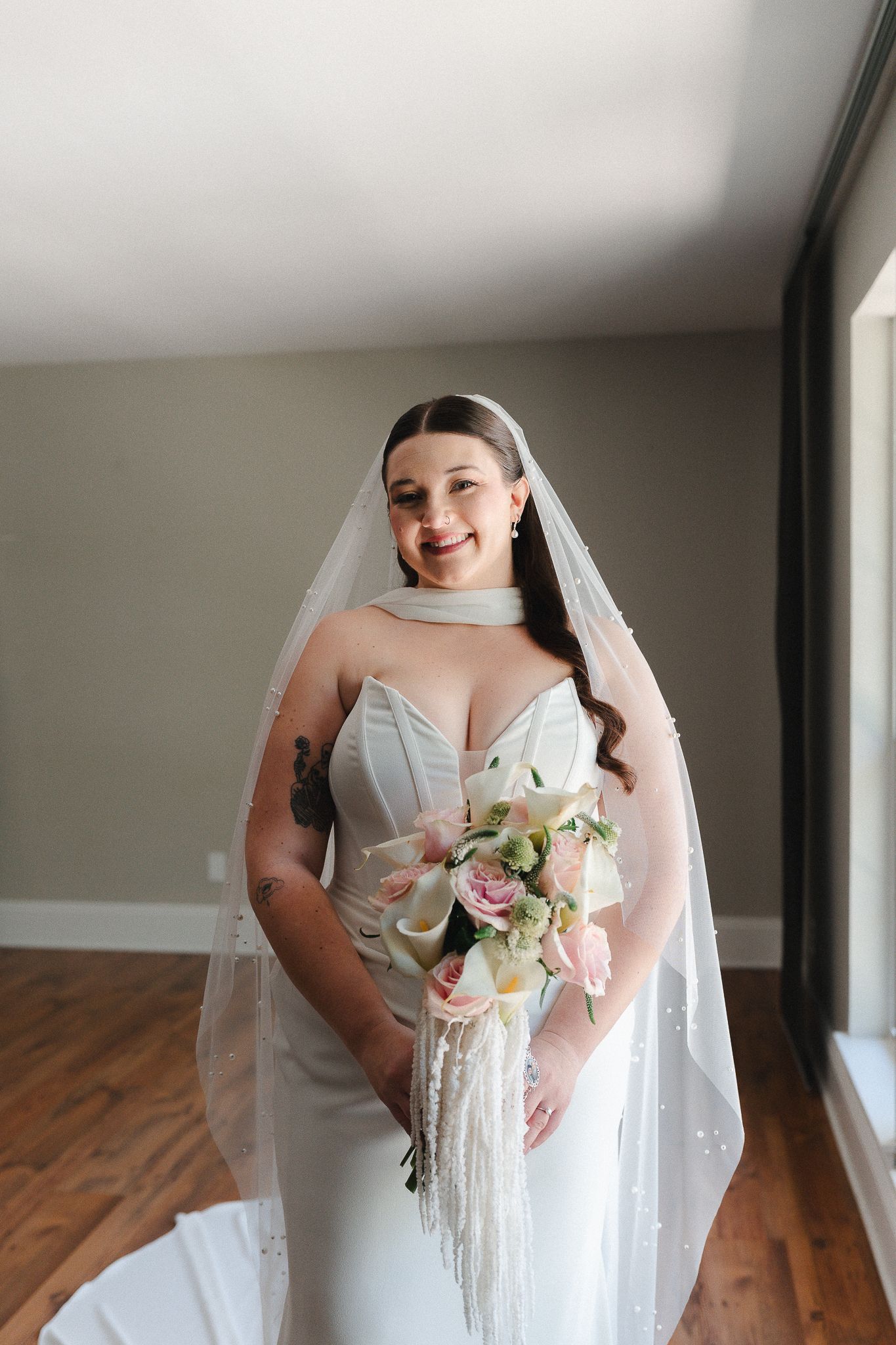 Smiling bride in white gown and veil holds pink floral bouquet with fringe accents indoors.