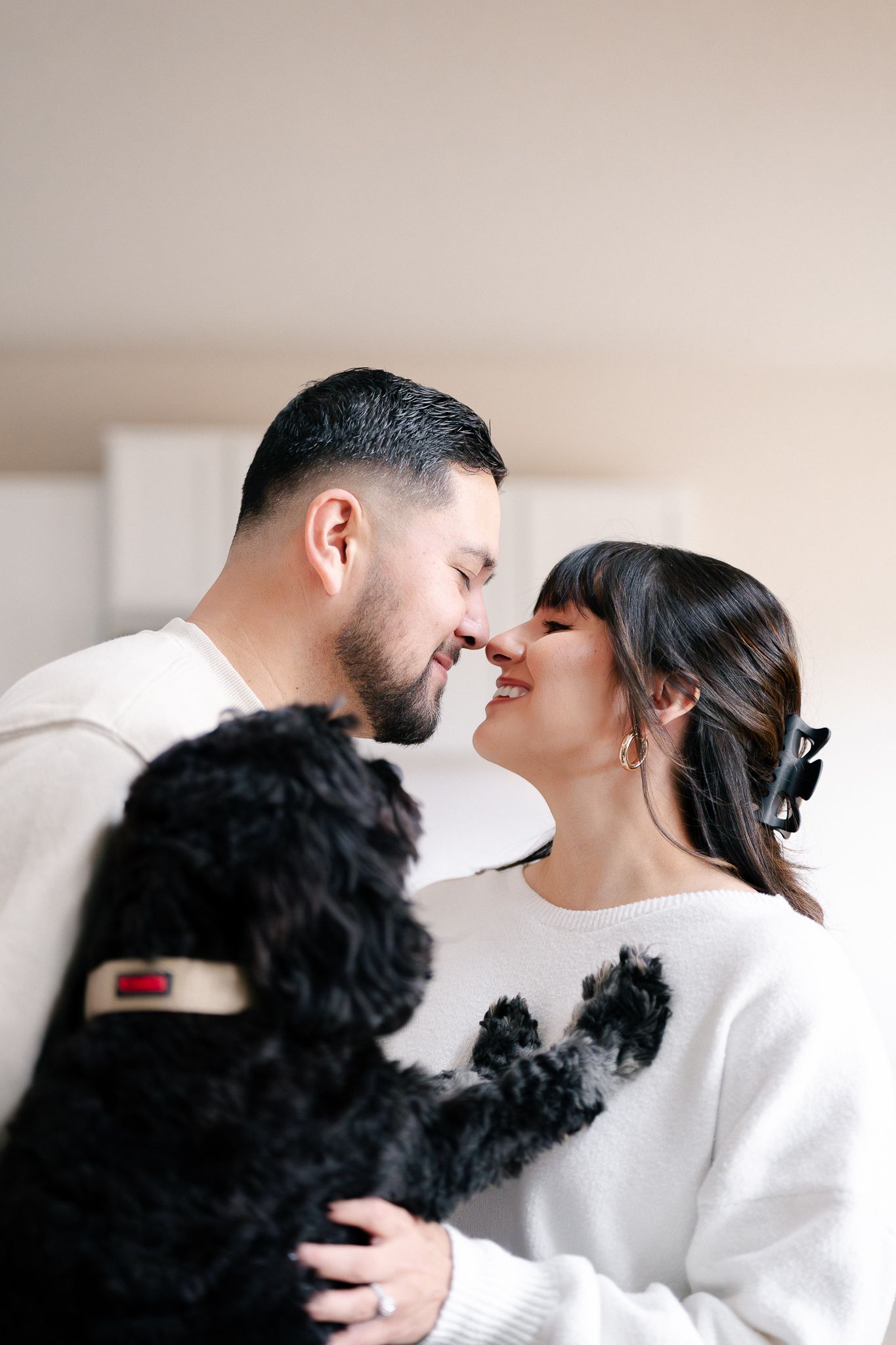 A couple in white sweaters shares tender moments while playing with their black dog in a bright indoor setting.