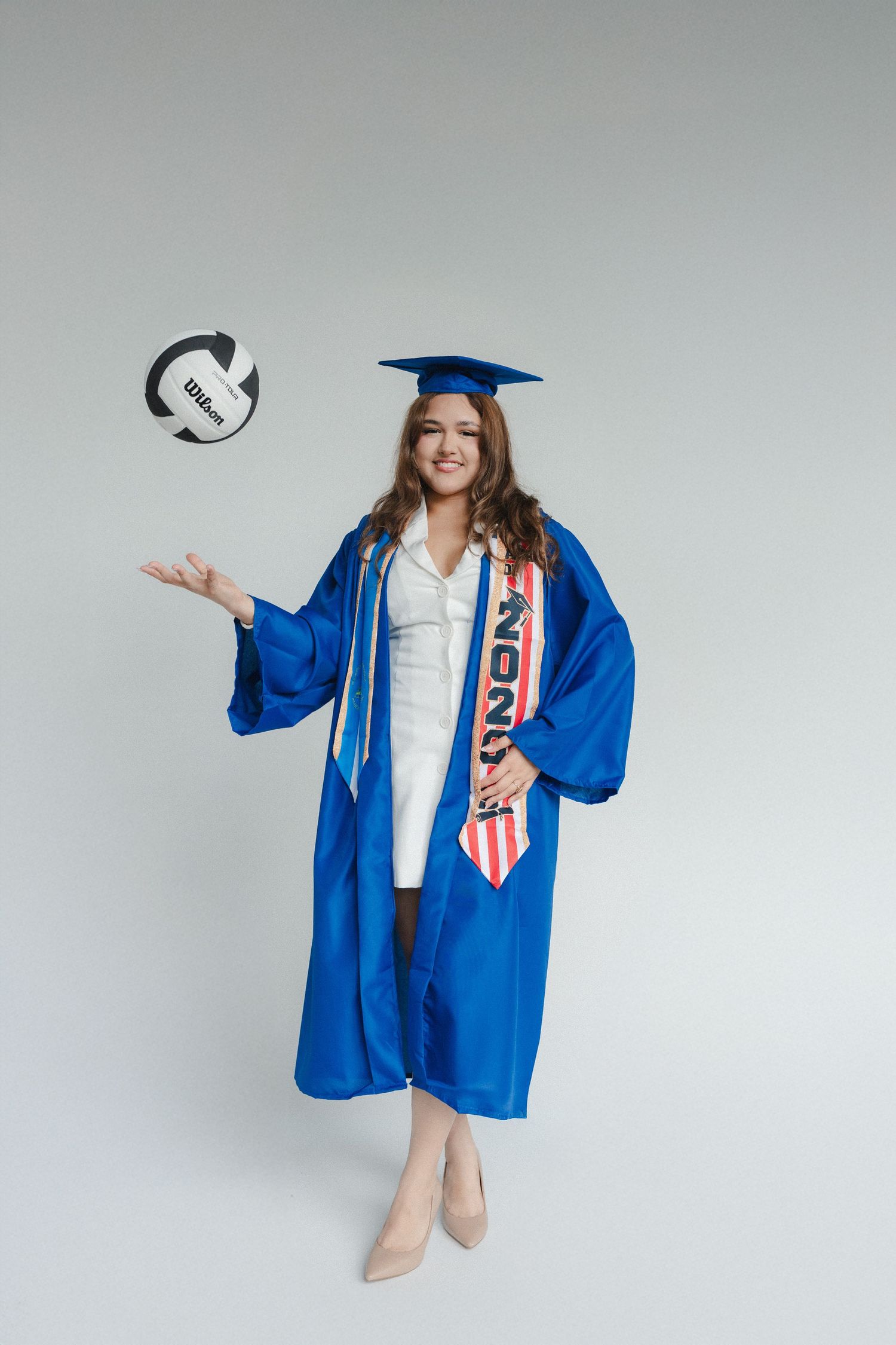 Young woman in blue graduation cap and gown tossing a volleyball, wearing heels and a sash, on white background.