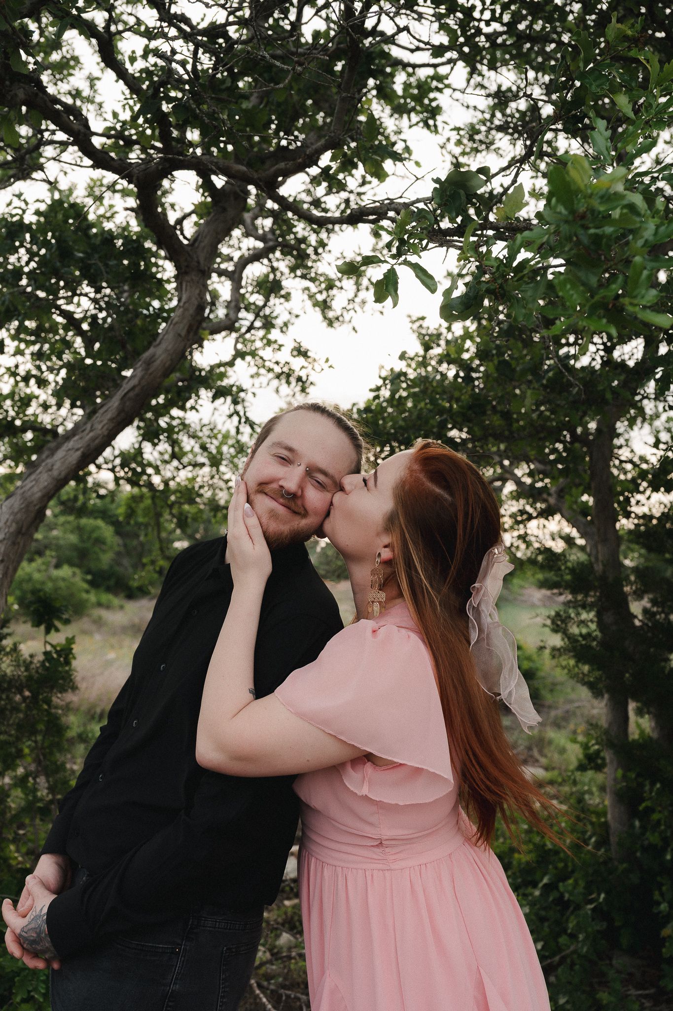 A woman in a pink dress kisses a smiling man in black jacket outdoors among lush green trees.