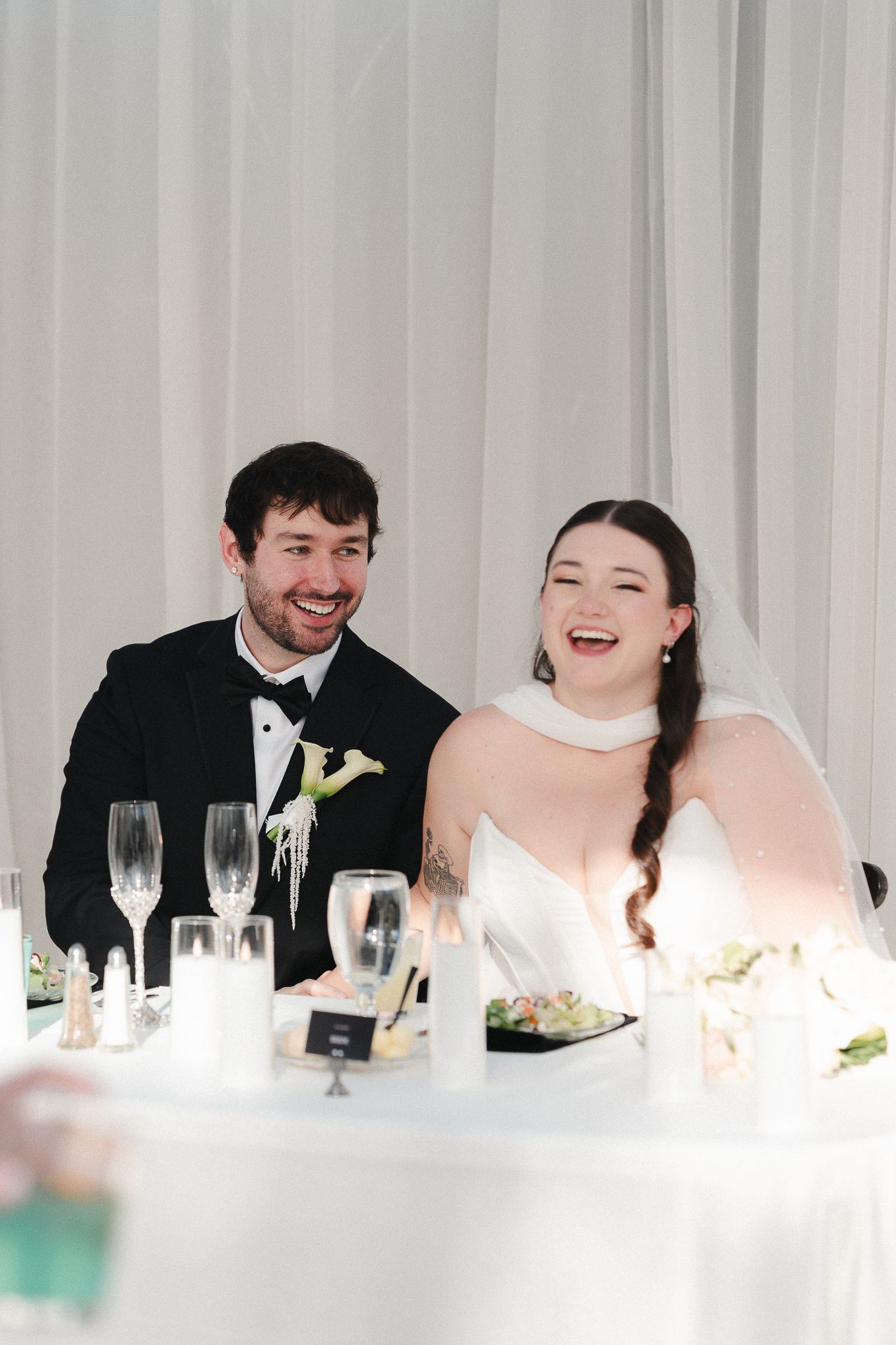 A joyful bride and groom laughing together at their wedding reception table, adorned with candles and champagne flutes.