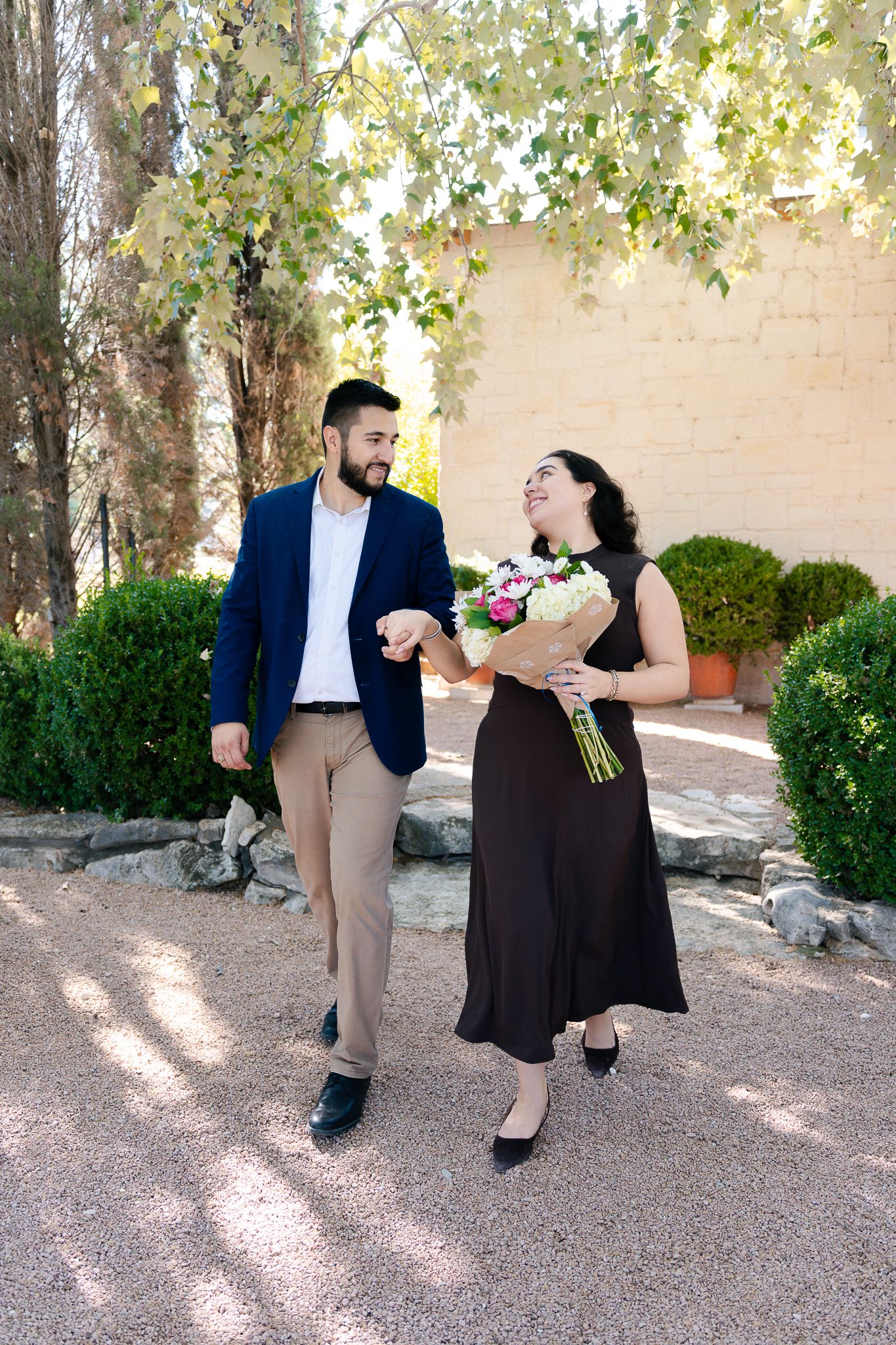 A couple in formal attire shares a romantic dance moment outdoors in a garden setting with dappled sunlight.