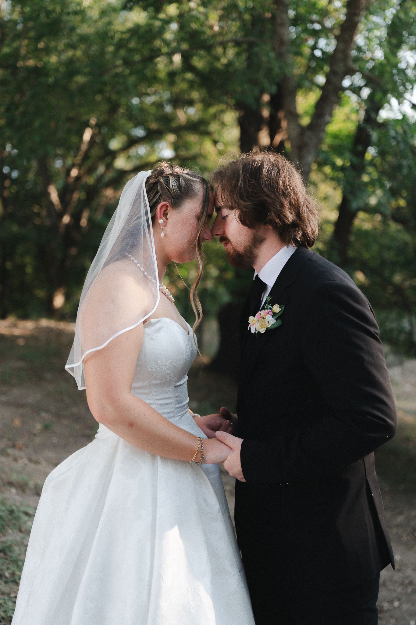 Bride in a white gown and veil touches foreheads with her groom in a dark suit and boutonniere during outdoor wedding portraits among trees.
