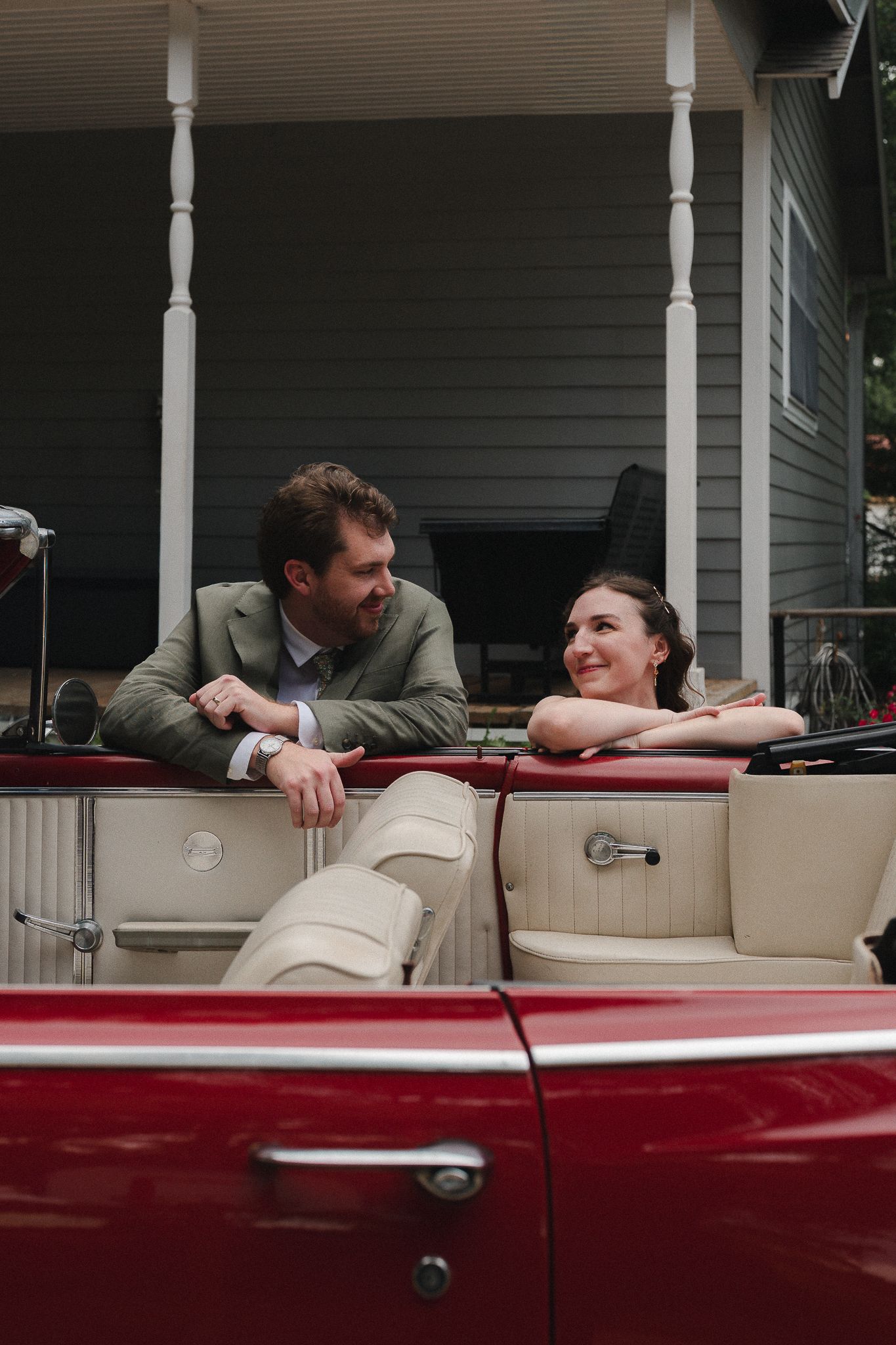 A couple in formal attire smiles at each other in a vintage red convertible car outside a house.