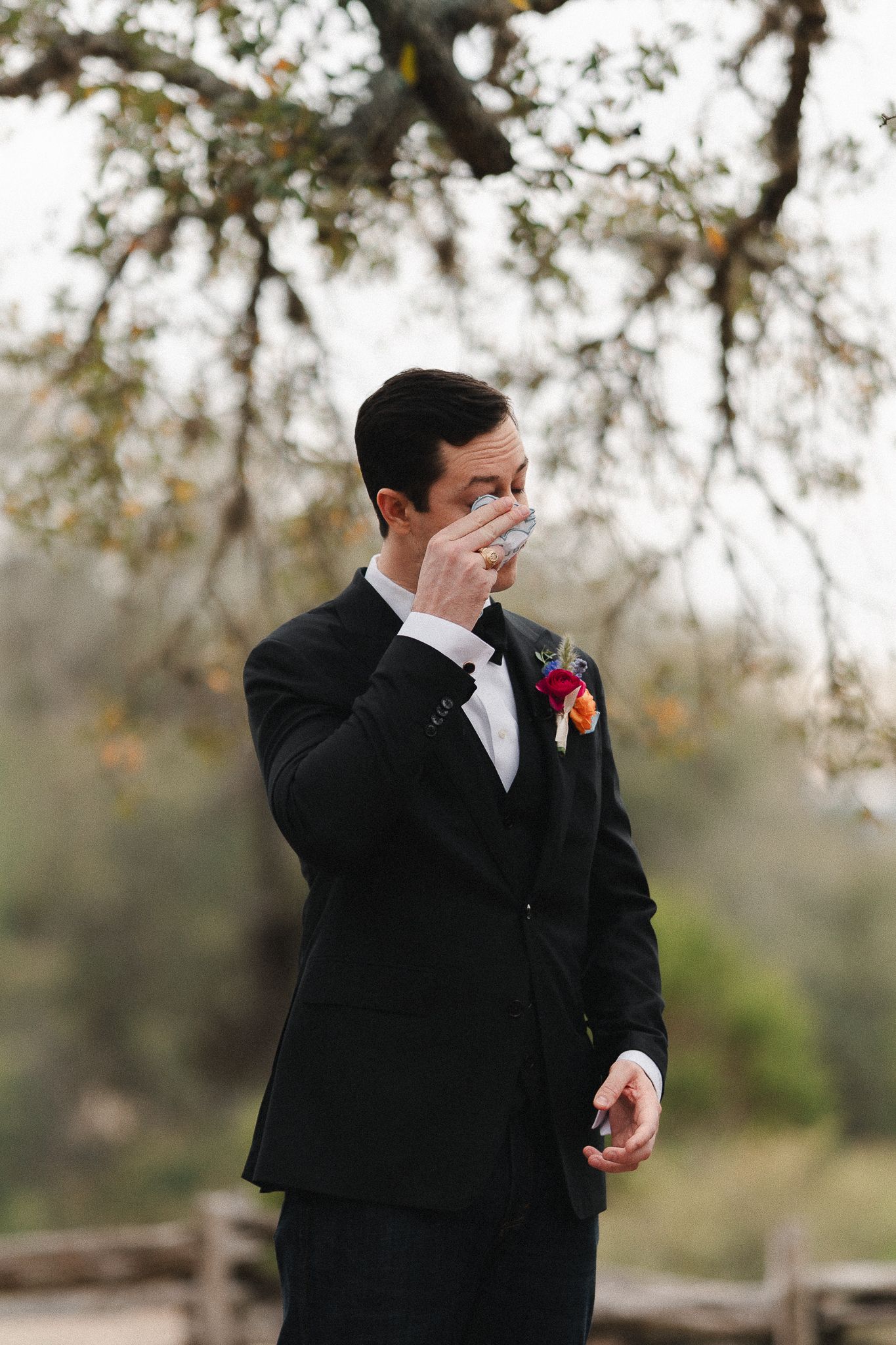 Emotional groom in black tuxedo wiping tears during wedding first look outdoors with floral boutonniere.