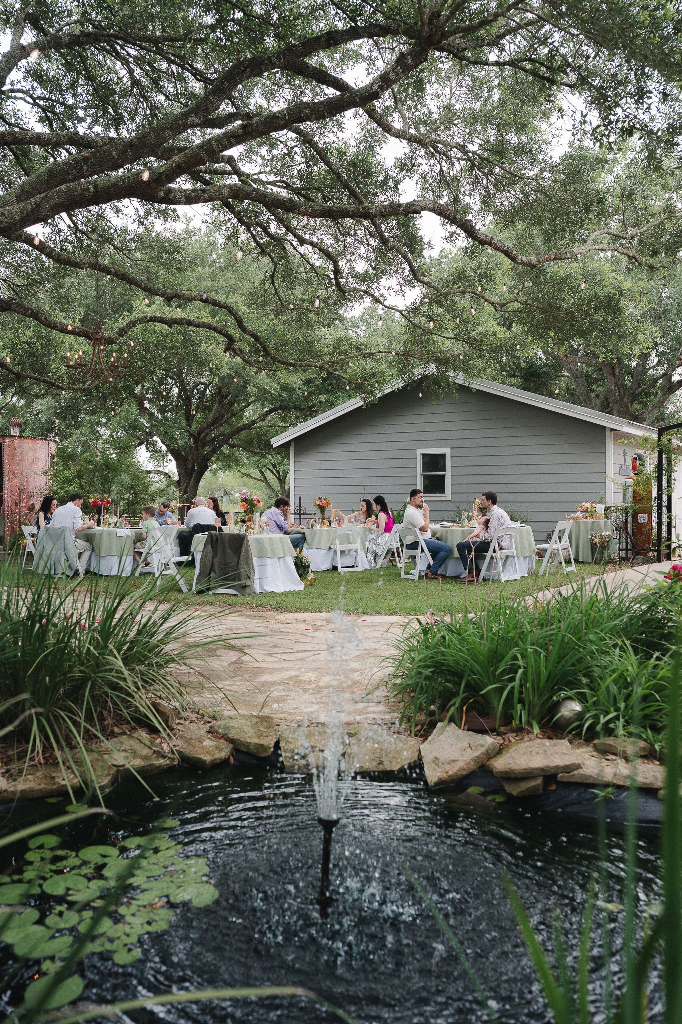 Outdoor wedding reception with guests seated at white tables near a fountain pond, surrounded by oak trees and a gray barn.