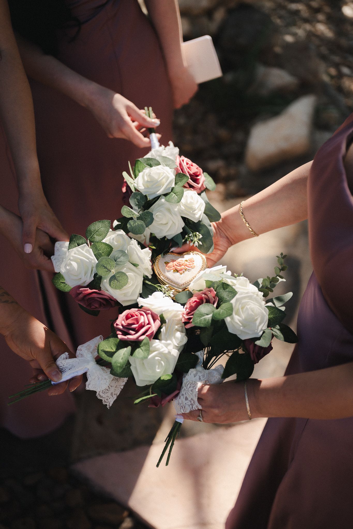 Bridesmaids in dusty rose dresses holding a bridal bouquet of white and burgundy roses with eucalyptus greenery.