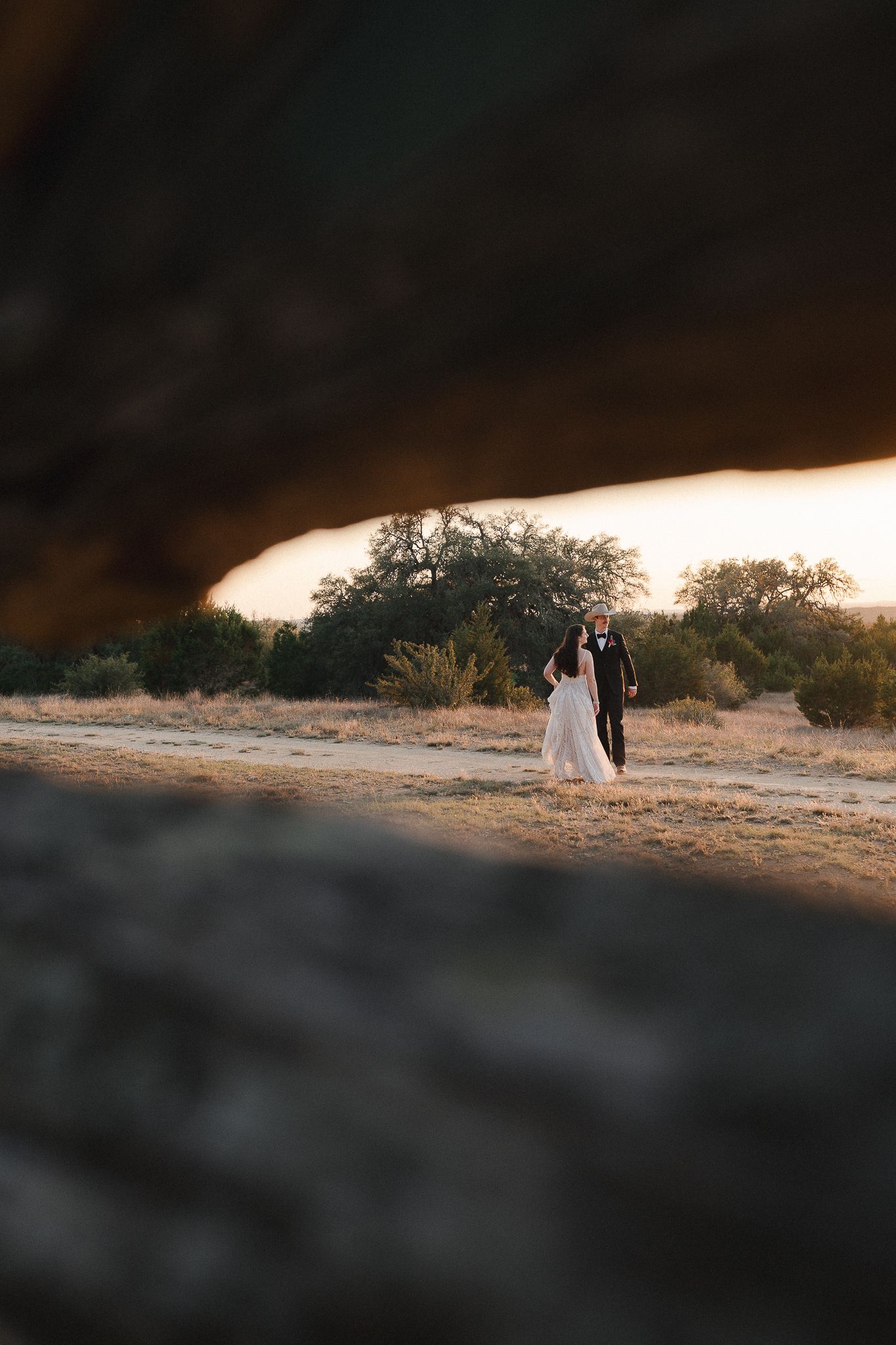 A bride and groom walk hand in hand through a desert landscape framed by rocky arch at golden hour.