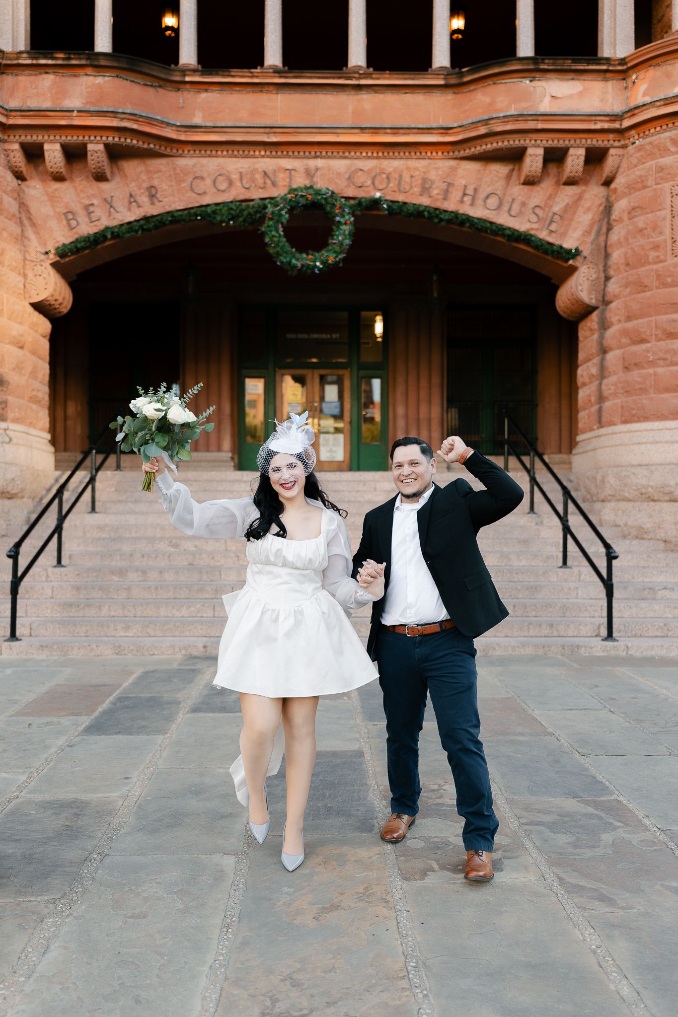 A couple in wedding attire playfully runs and jumps through a brick archway in a courthouse building.