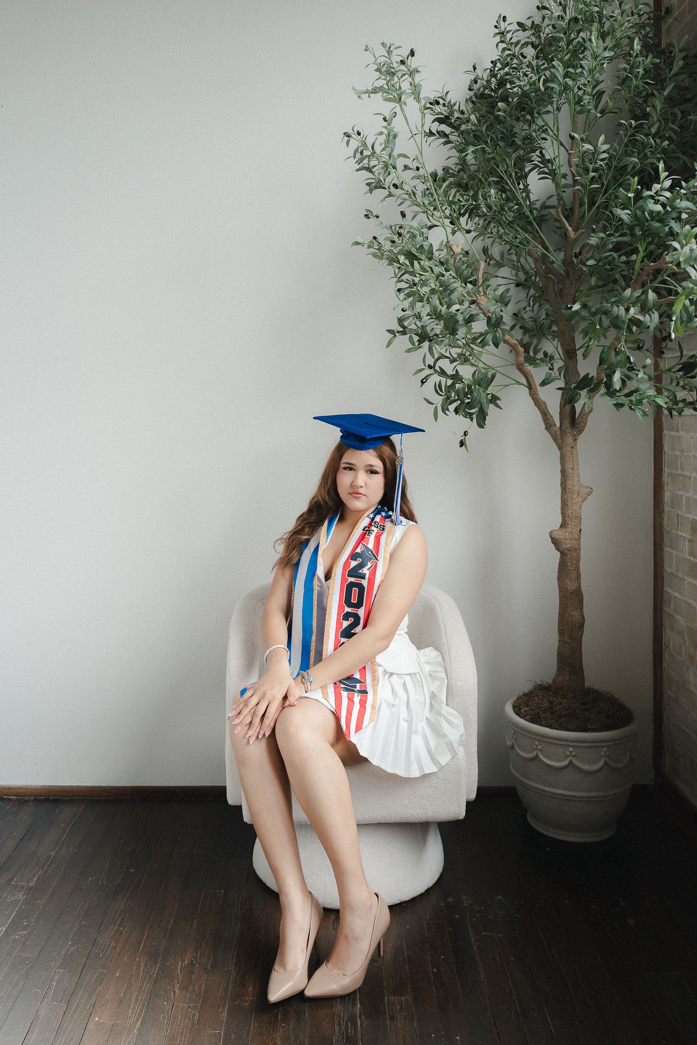 Young woman in graduation cap and sash seated in white chair beside indoor tree, celebrating class of 2023.