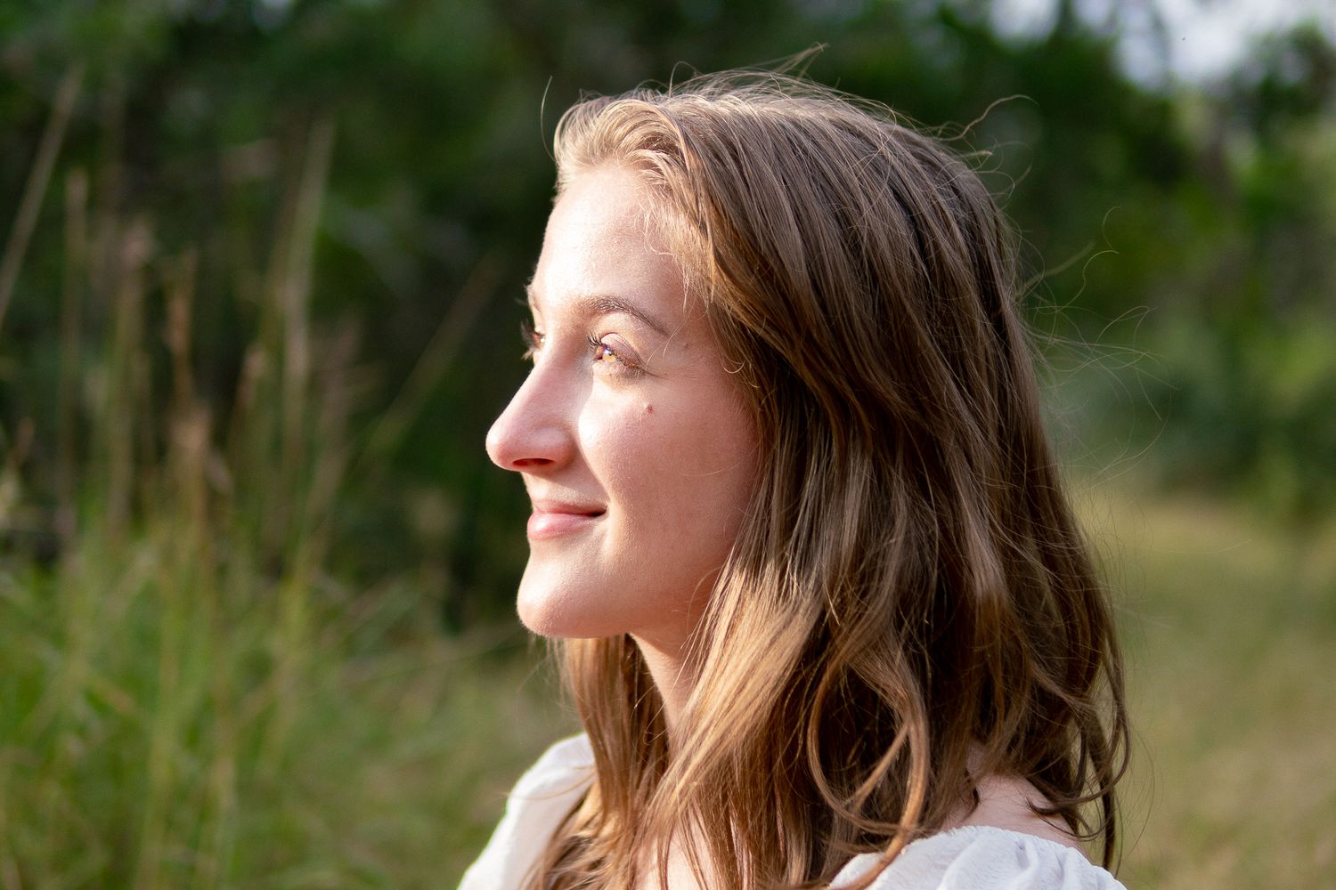 A person in white clothing laughs while standing in a natural outdoor setting with green foliage in the background.