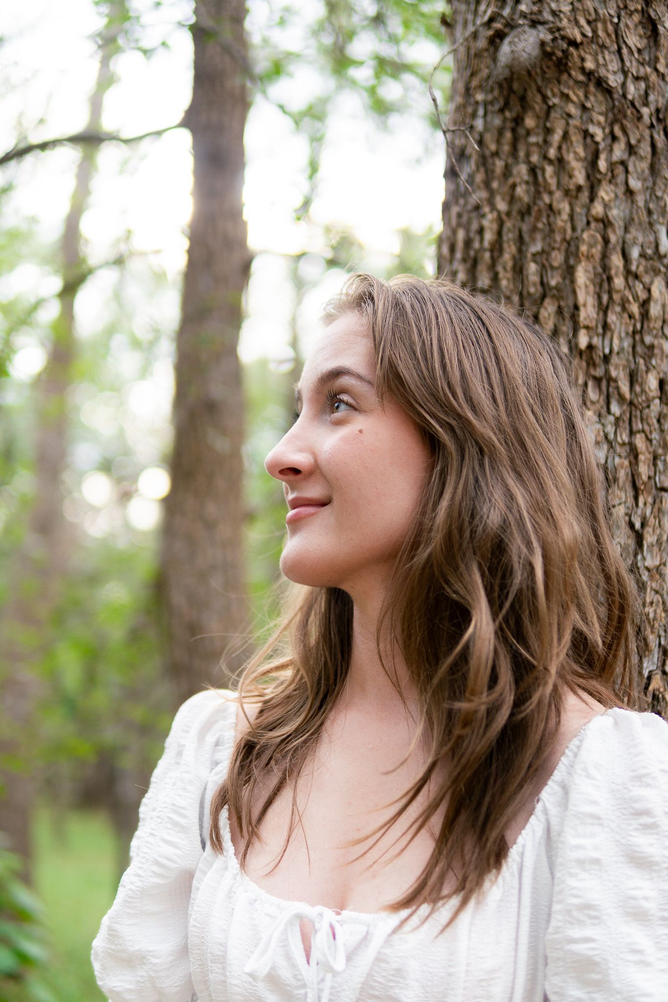 A woman in a white blouse looks off to the side while standing in a forest setting with trees in the background.