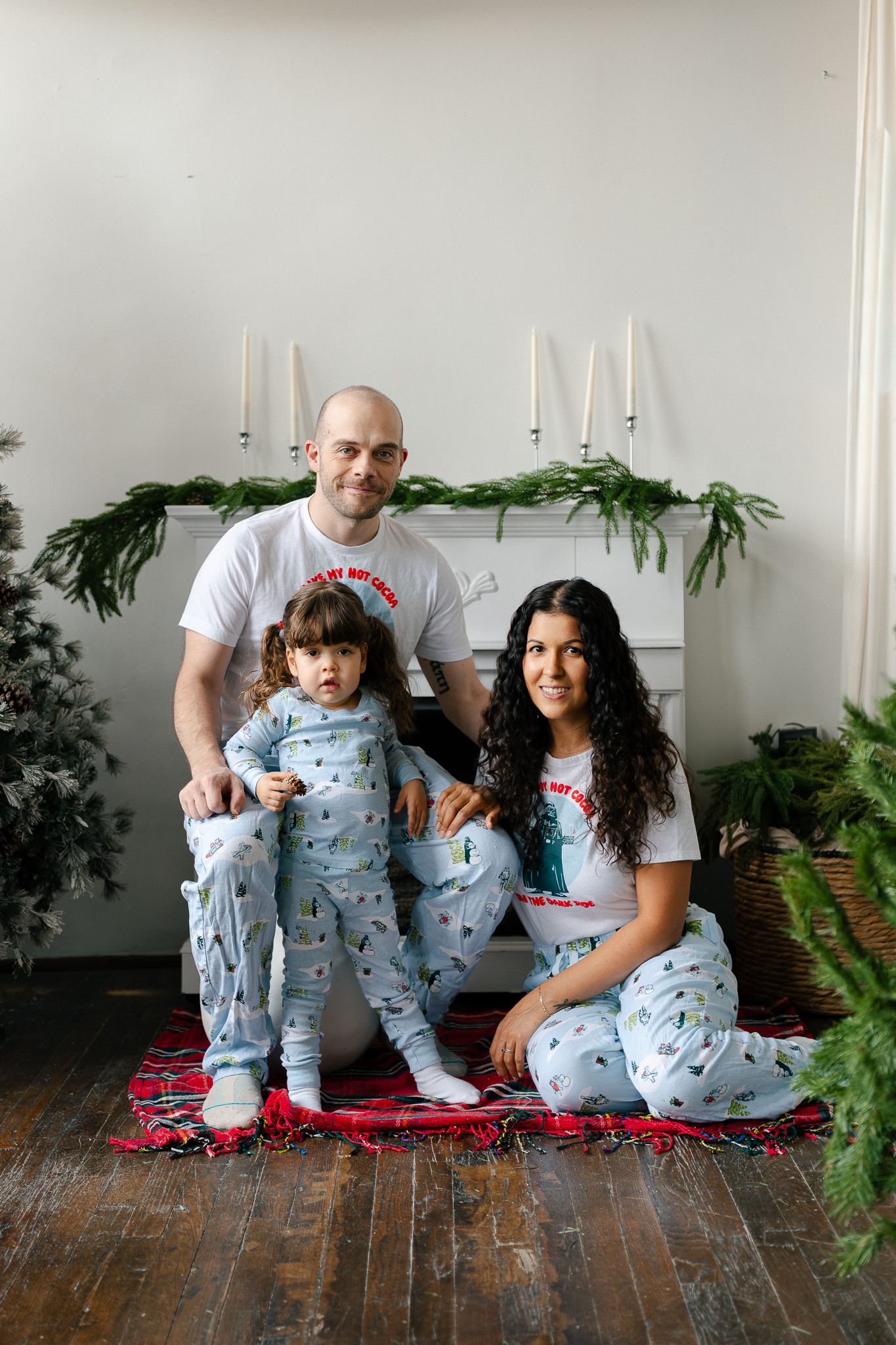 A family wearing matching heart-print pajamas sits together on a wooden floor with plants in the background.