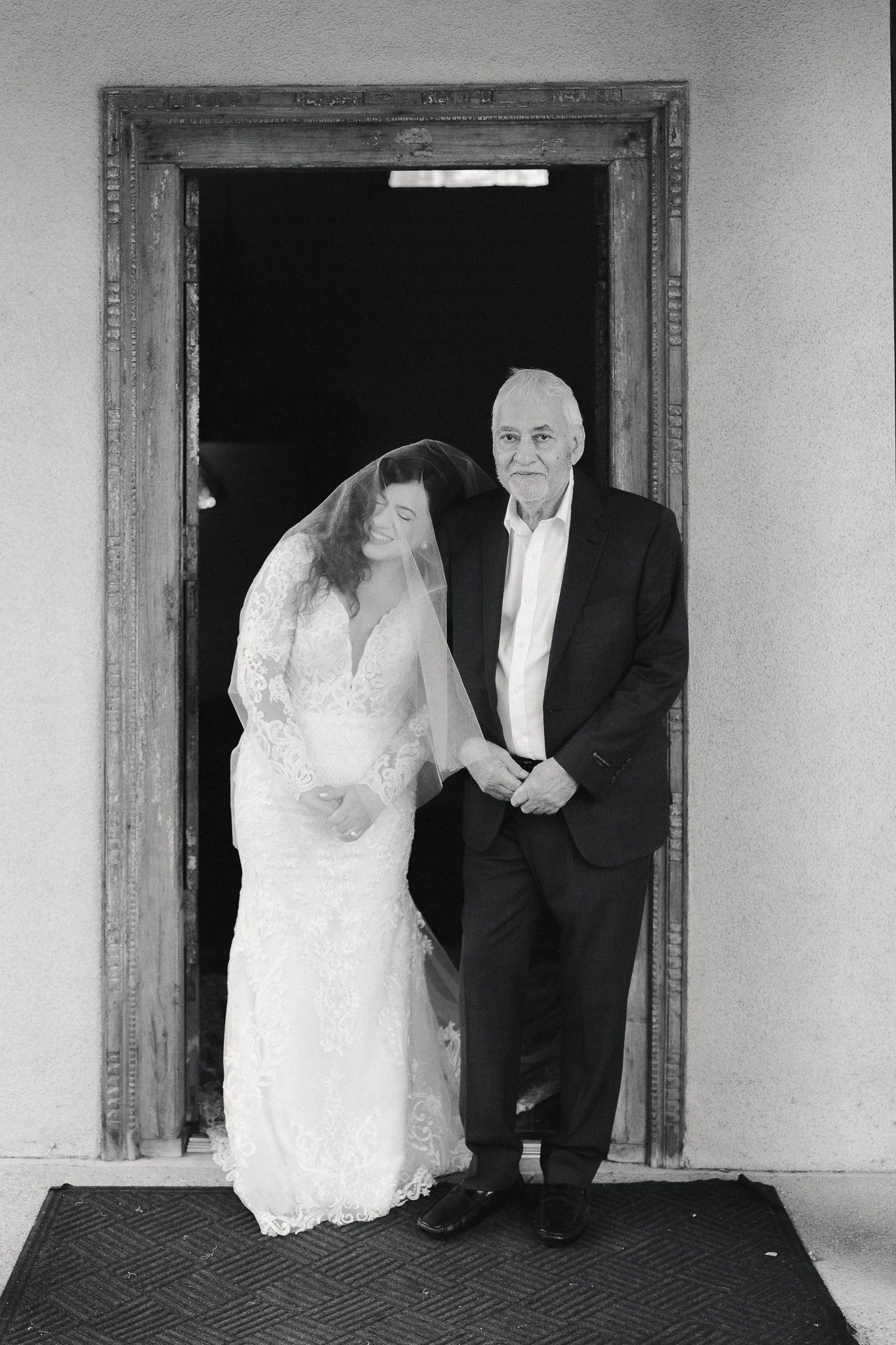 Black and white photo of a bride in a lace gown and veil standing beside an older man in a suit in a rustic wooden doorway.