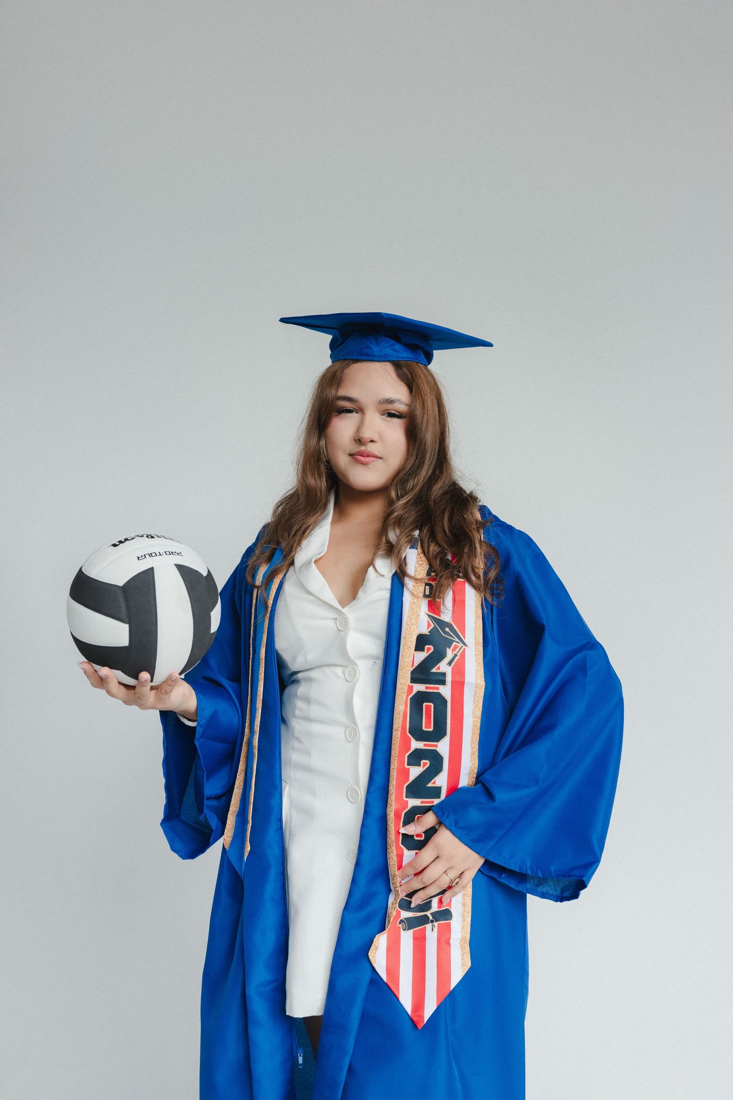 Graduate in blue cap and gown holds volleyball, wearing a 2023 sash, posing for senior portrait.