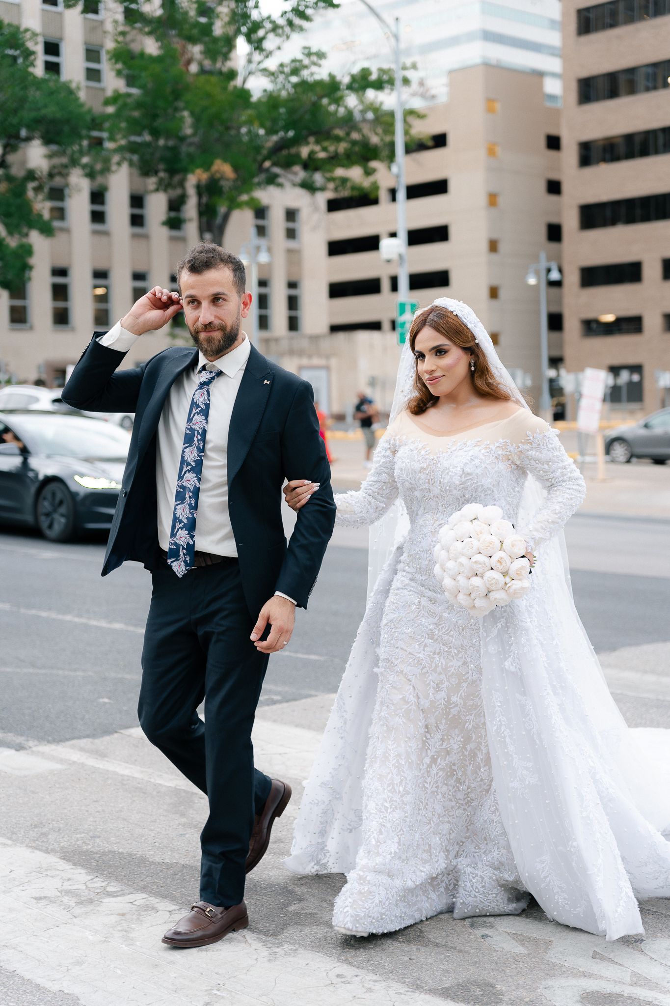 A couple in wedding attire playfully poses in a parking lot with tall buildings in the background.