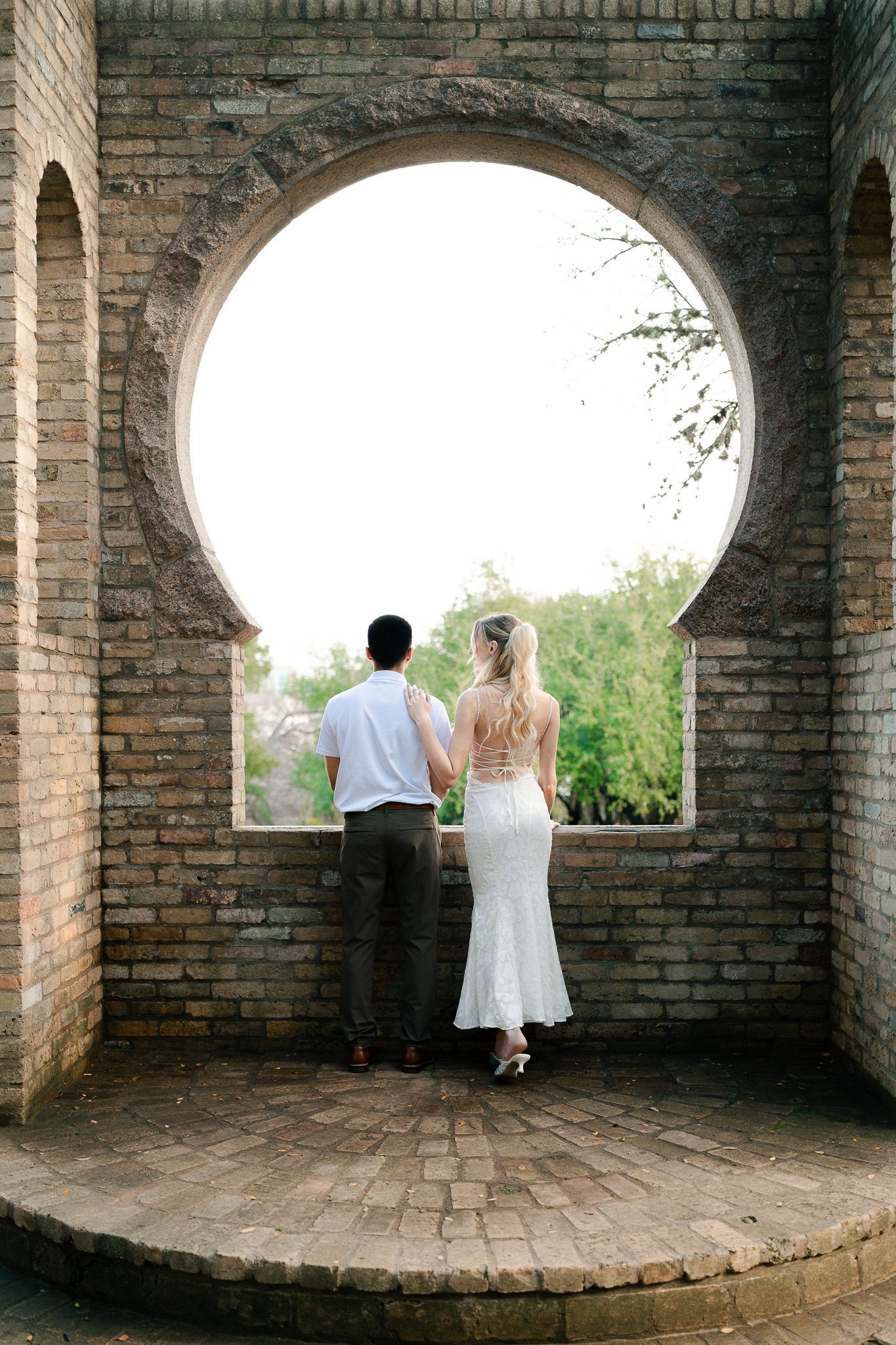 Couple in formal attire stands in arched brick passageway overlooking scenic garden view.