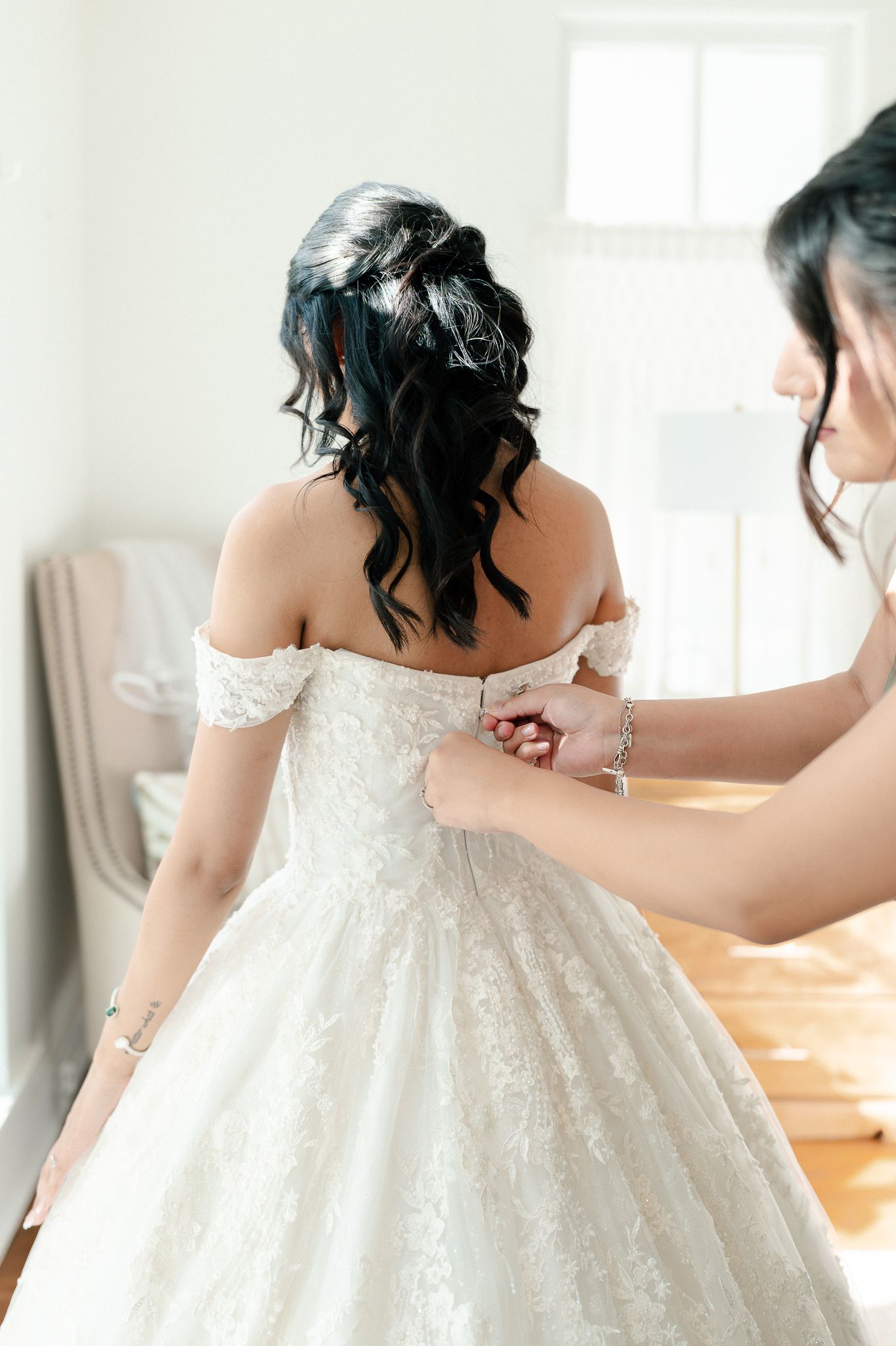 A bride in a white lace wedding dress being helped with final adjustments to her gown before the ceremony.