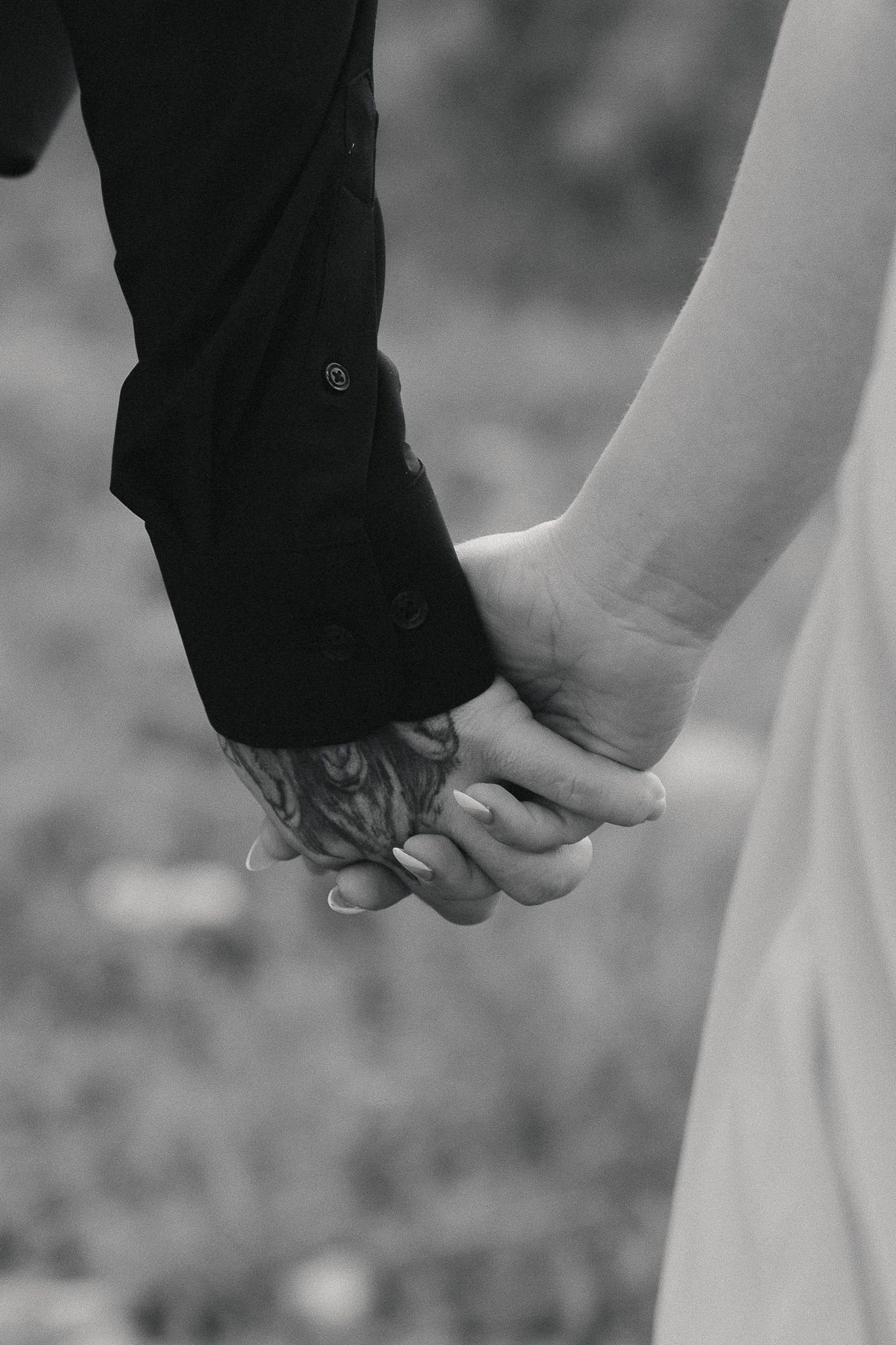 Black and white photo of a couple holding hands, groom with tattoo in dark suit, bride in white dress.