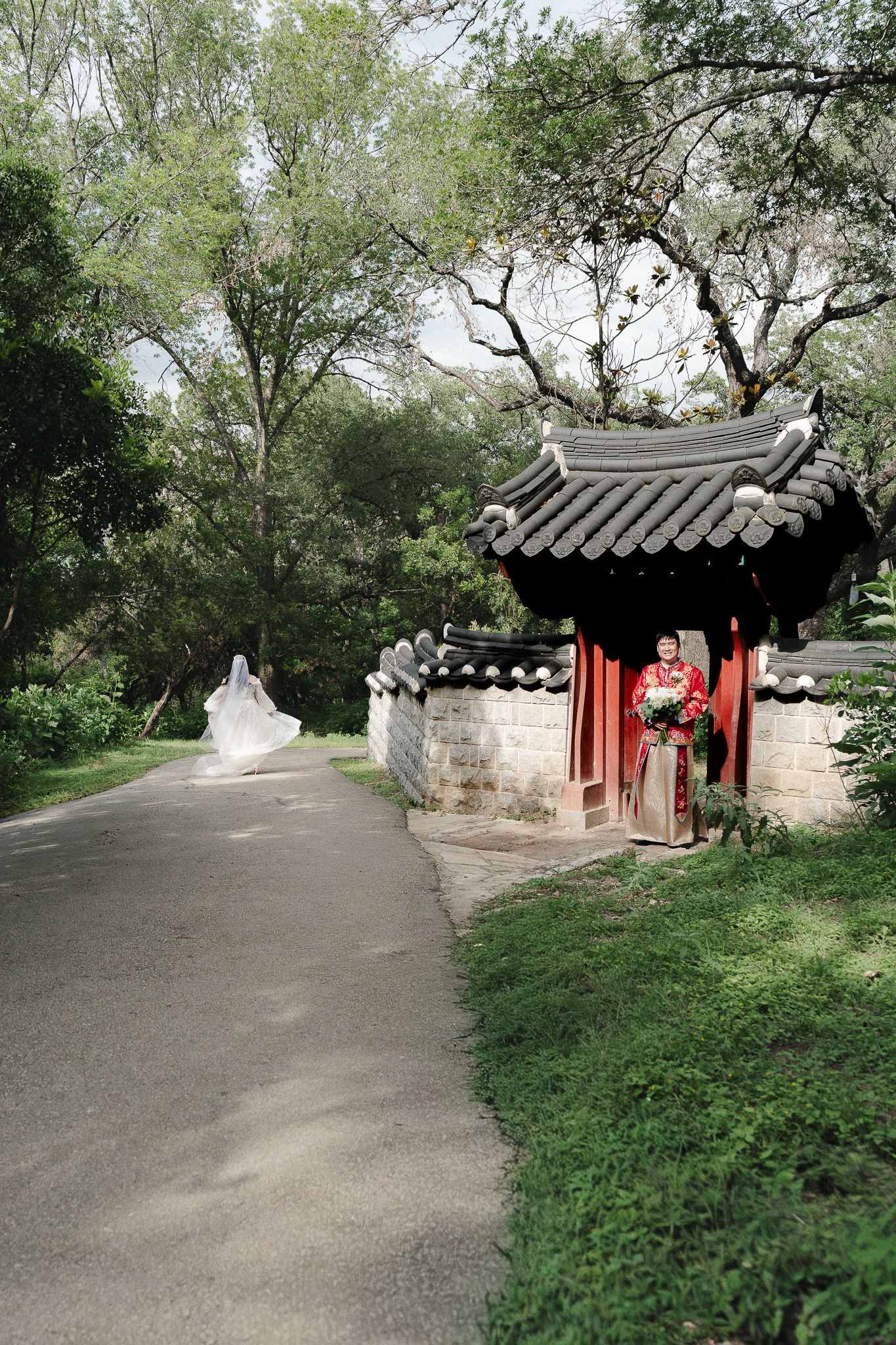 Bride in white gown and groom in red traditional attire pose near a Chinese garden pavilion surrounded by lush trees.
