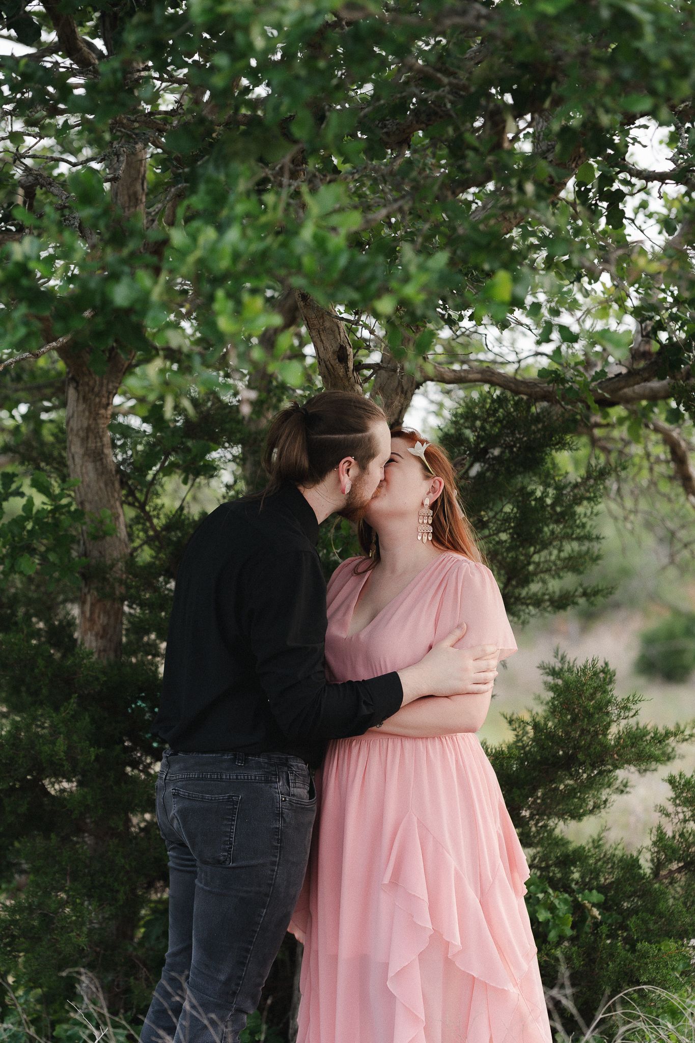 Couple sharing a romantic kiss outdoors, woman in pink dress embraced by man in black, surrounded by lush green trees.