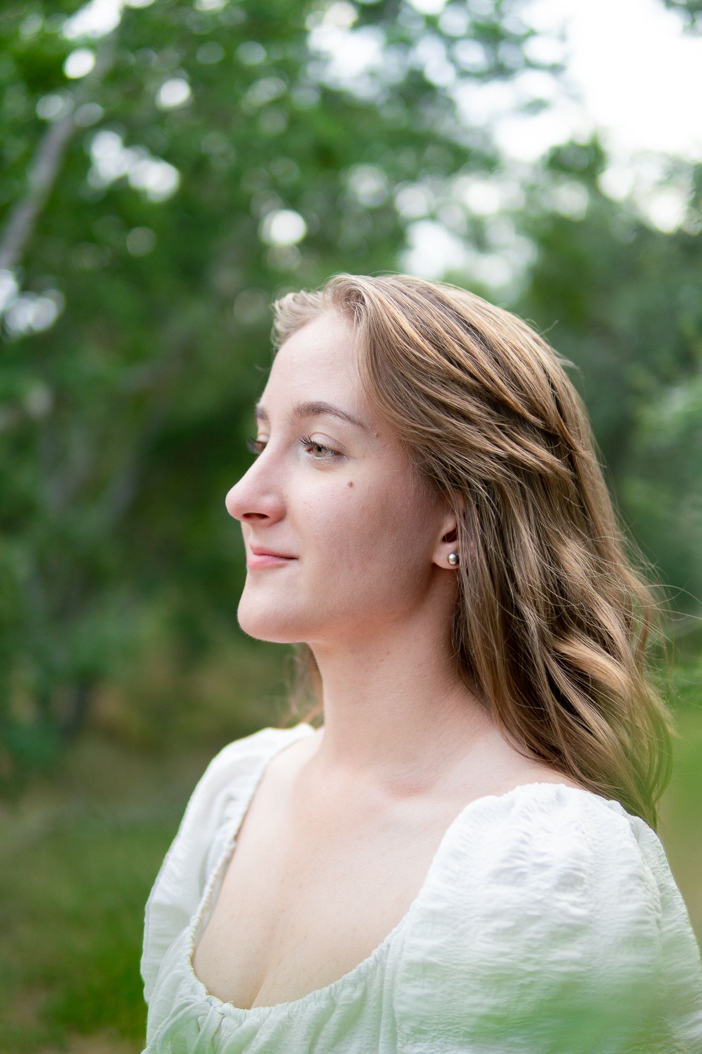 A person in a white dress gazes off to the side with soft natural lighting and green foliage in the background.
