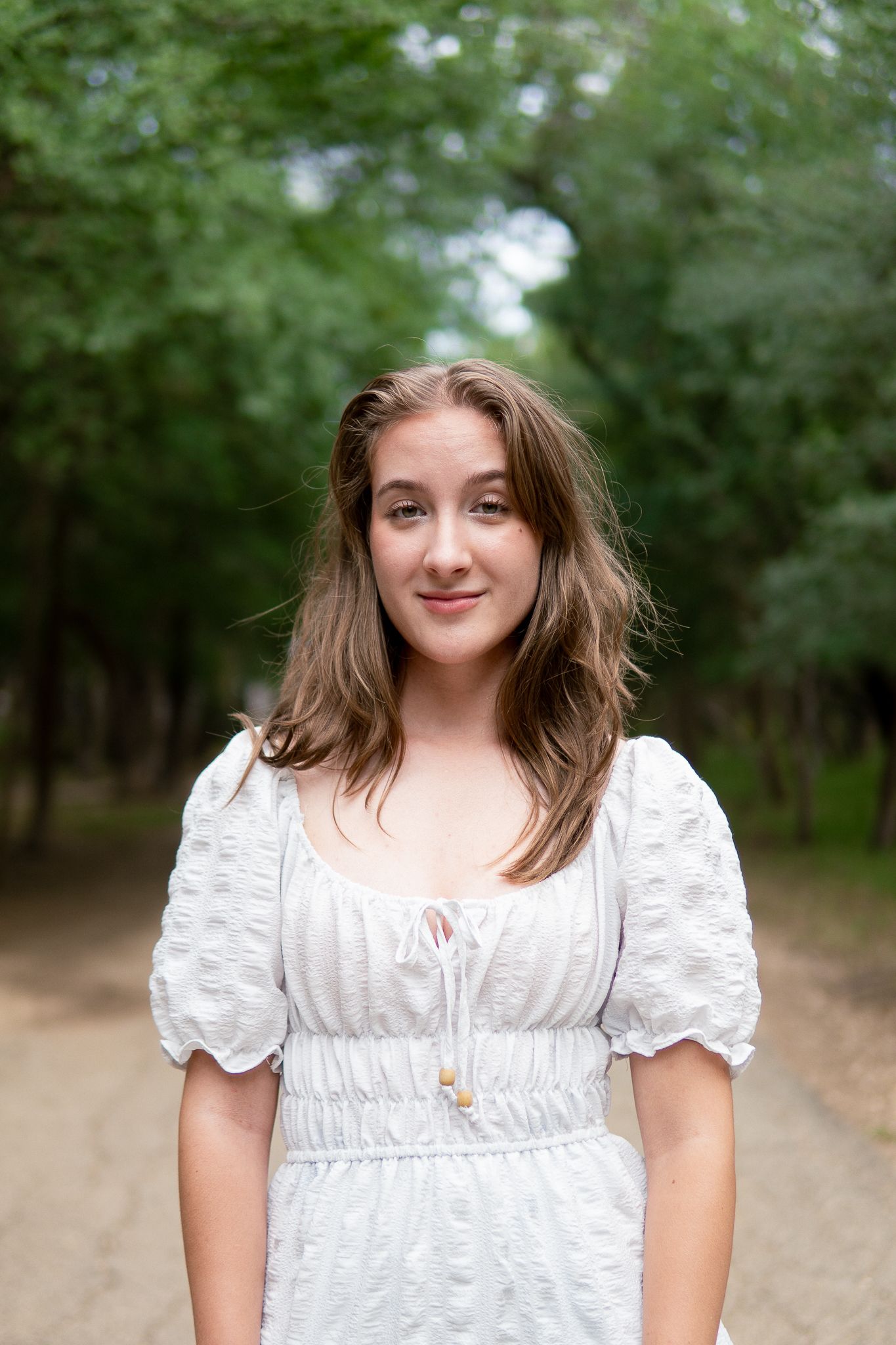 A person in a white puffy-sleeved dress smiles while standing outdoors in a wooded setting.