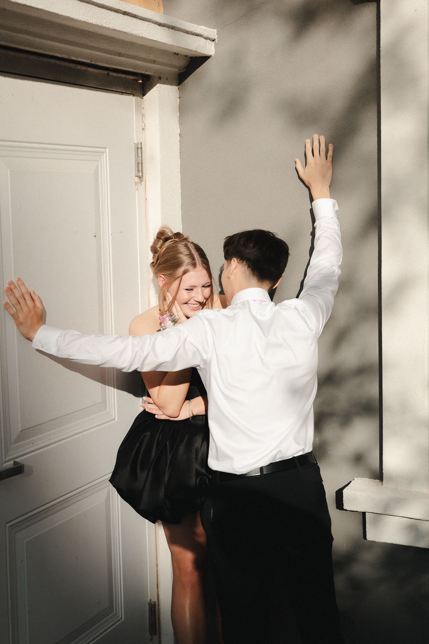 Couple dancing joyfully outside a white door, woman in black skirt and man in white shirt with arms raised.