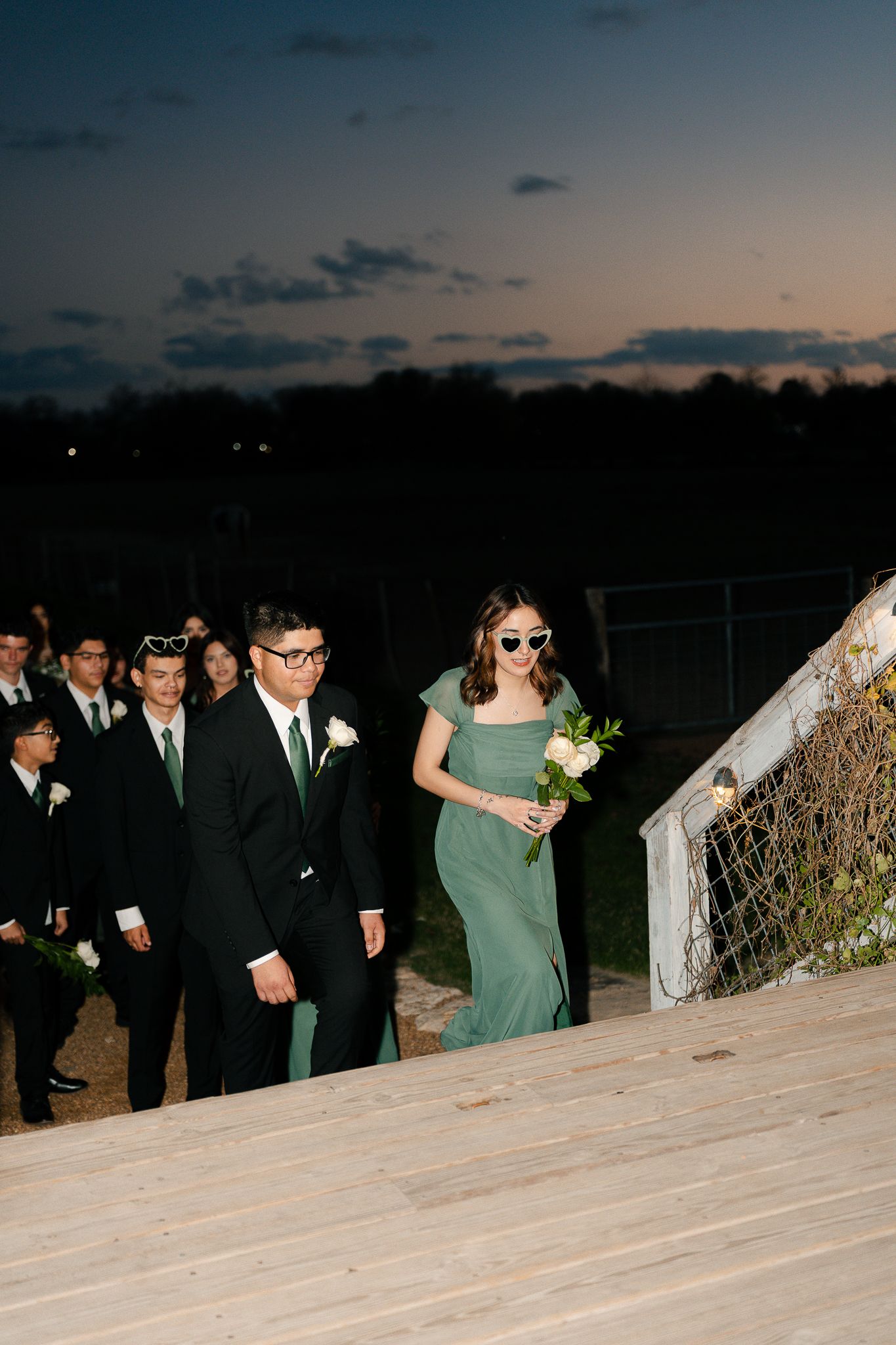 Wedding party members walk along a stone wall at dusk during an outdoor celebration.
