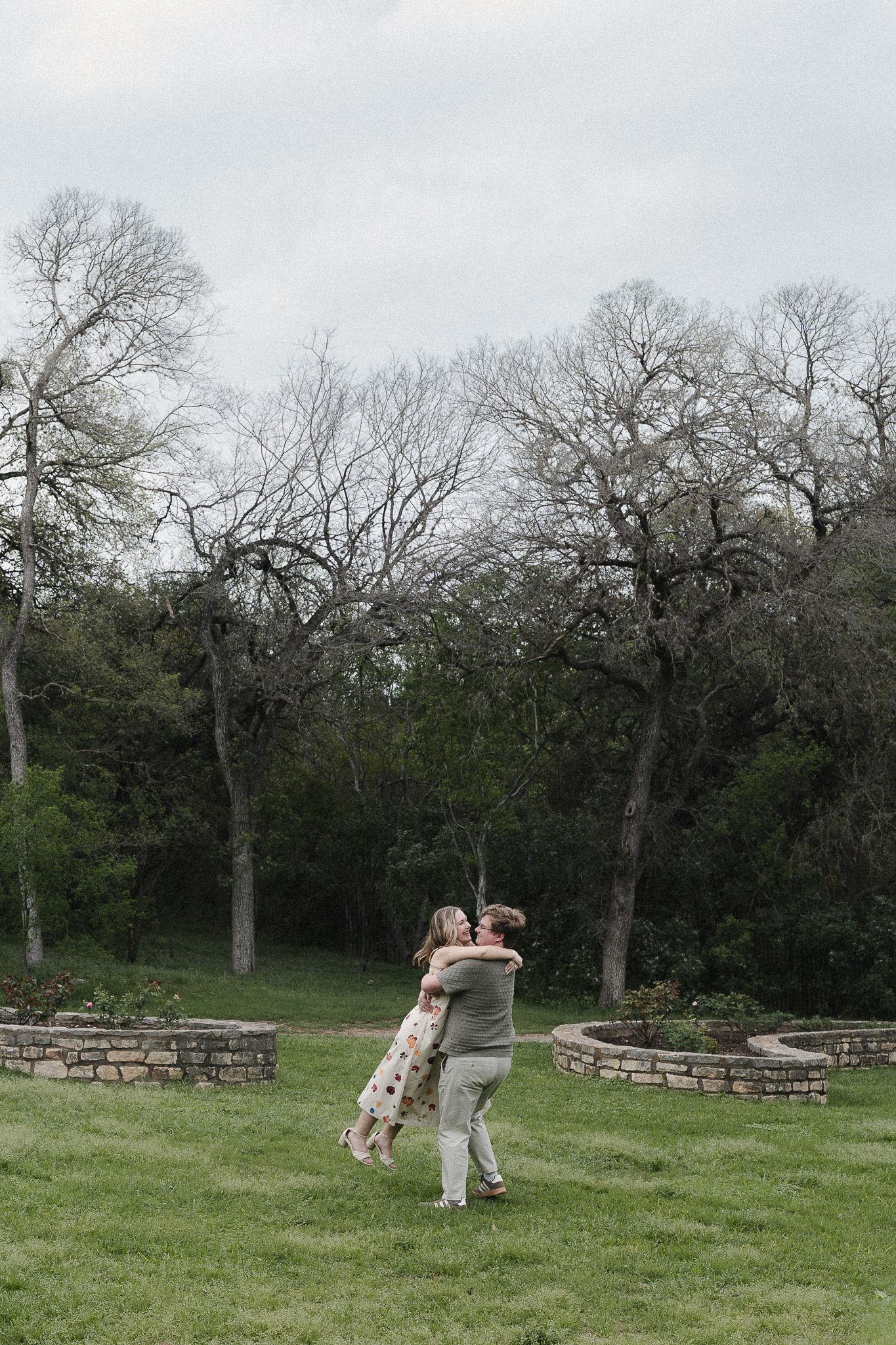 Couple embracing and spinning on green grass surrounded by bare trees and stone garden walls on a cloudy day.