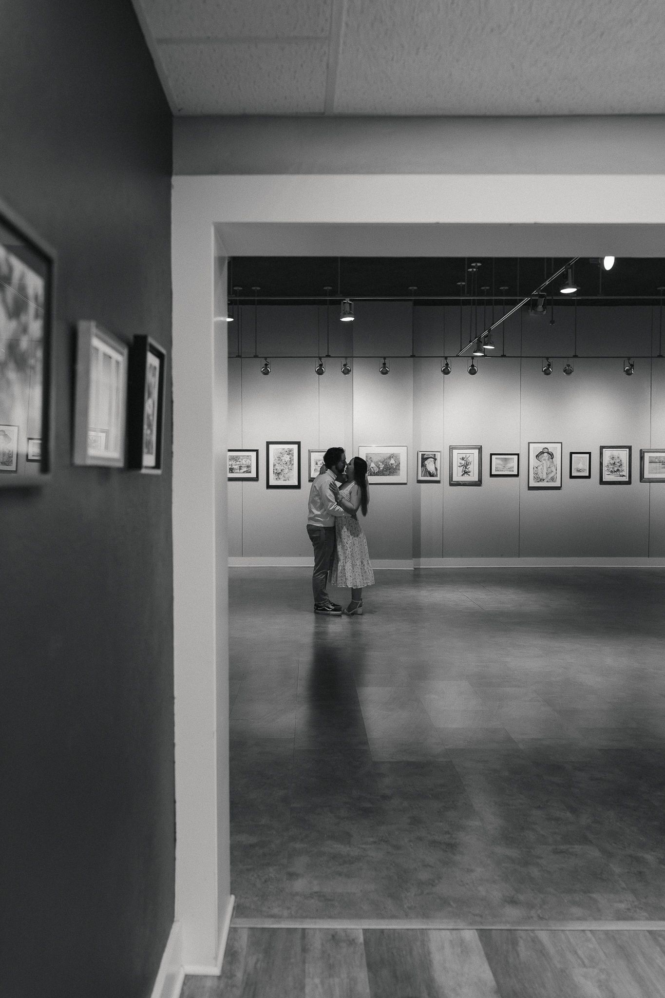 Black and white photo of a couple embracing in an art gallery with framed artwork on walls.