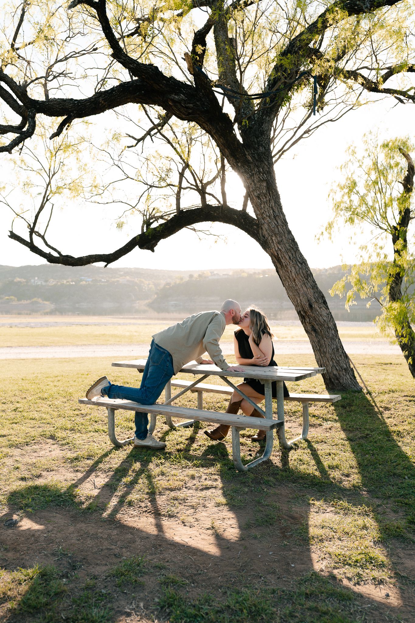 A couple shares a tender moment at a picnic table under a large tree during golden hour at a park.