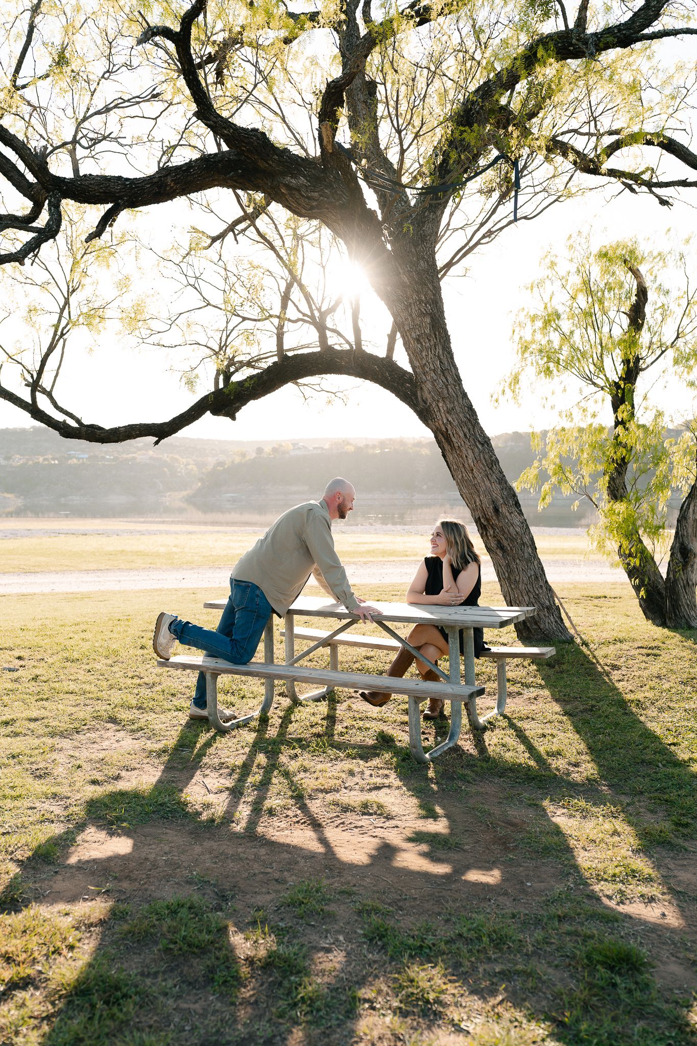 Couple sharing a moment at a picnic table under a sunlit tree in a peaceful outdoor park setting.