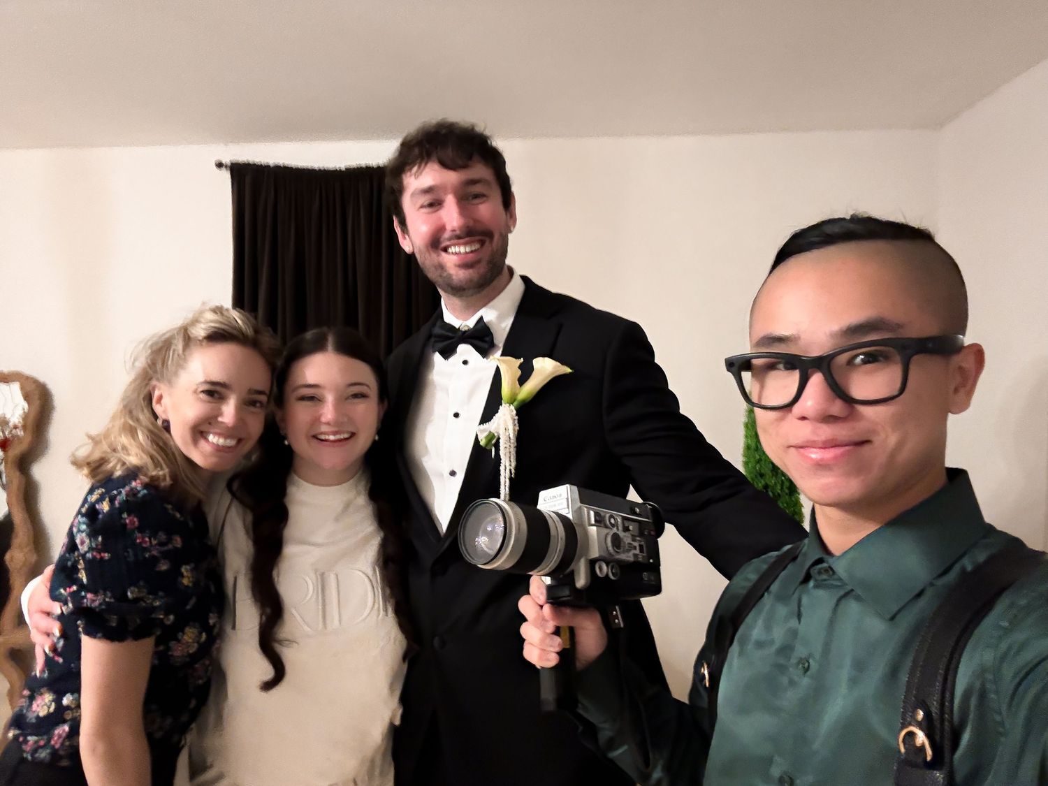 Four young people smiling for a selfie, one wearing a tuxedo with boutonniere and holding a camera.