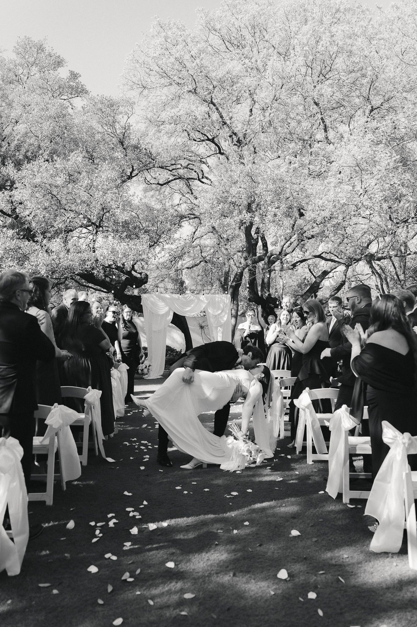 Black and white photo of a bride and groom sharing a kiss at the altar during an outdoor wedding ceremony.